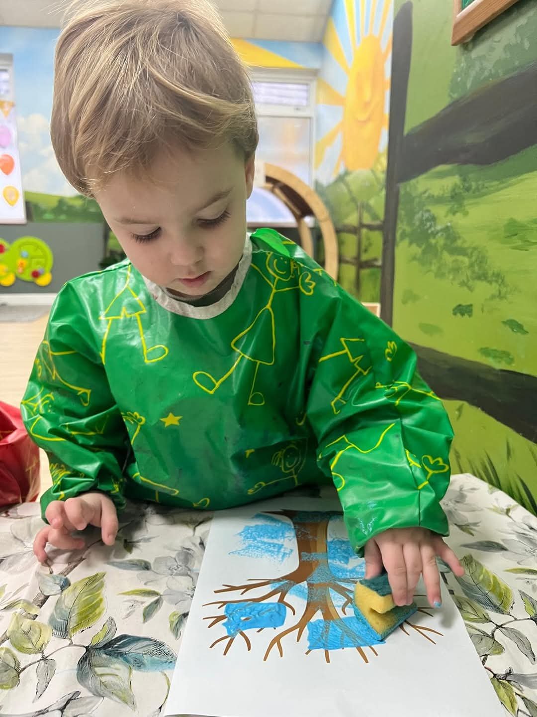 A young boy in a green apron is coloring a tree drawing with a sponge, in a colorful classroom with a nature mural on the wall.