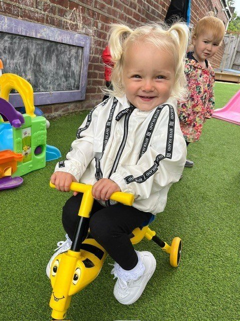 Toddlers playing on artificial grass with one child riding a yellow toy bike and another child in the background wearing floral clothing. Outdoor play area with colorful play structures visible.