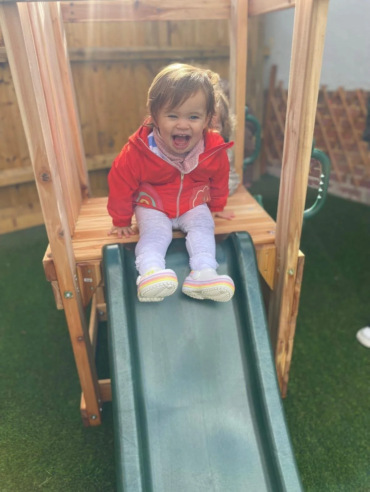 A young girl with brown hair sitting at the top of a small green slide, smiling and laughing, wearing a red jacket and light gray pants, on a playground with a wooden structure and green turf.