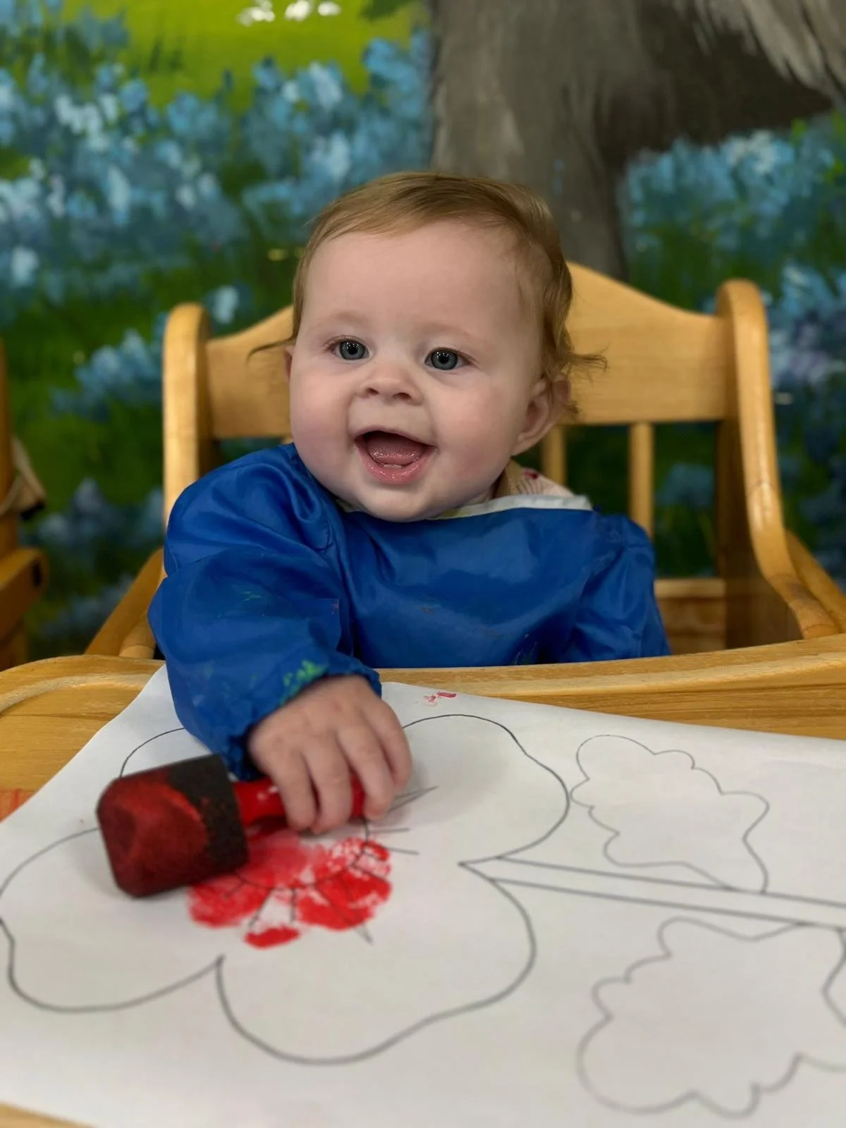 A cheerful baby with light brown hair and blue eyes, wearing a blue smock, is sitting at a wooden table coloring with a red crayon. There is a coloring sheet with outlines of flowers on the table. The background features a nature scene with trees and