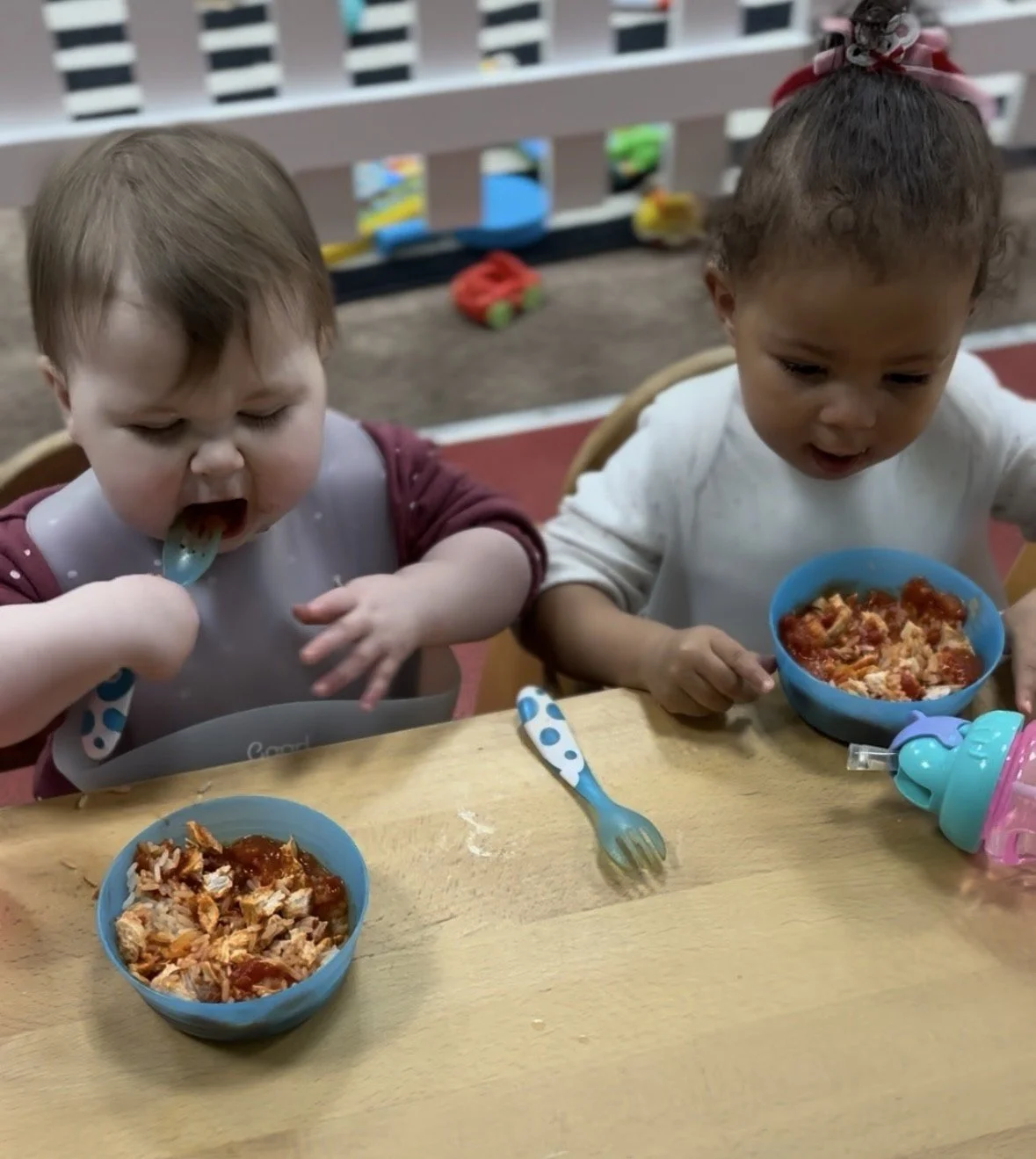Two young children sitting at a table, eating bowls of spaghetti with tomato sauce. The child on the left with a bowl in front has a spoon in their mouth, and the child on the right is holding a spoon near their bowl. There are some utensils and a sippy cup on the table.