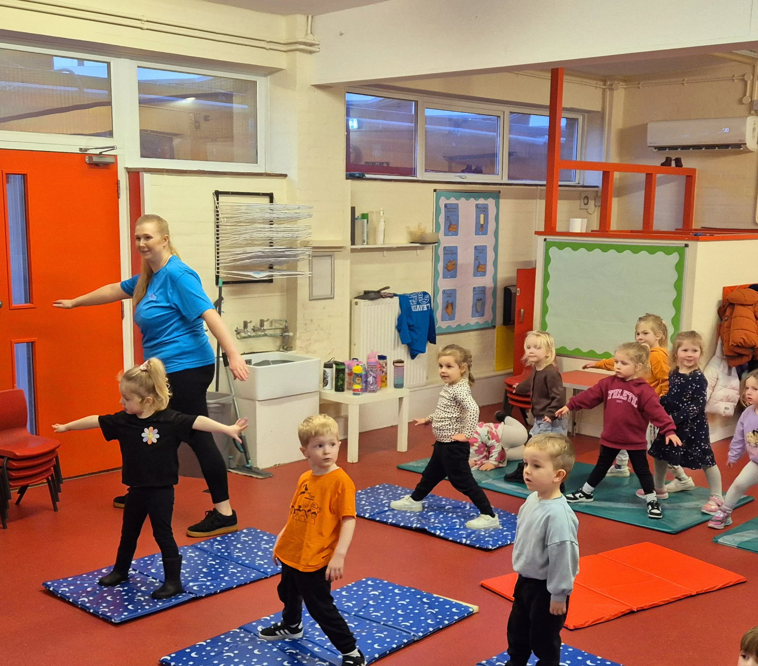 Children participating in a classroom activity, standing on mats, with a teacher leading the group.