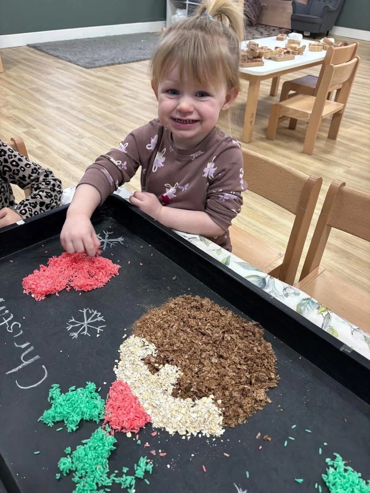 A young girl with red hair and a brown shirt decorated with white bunnies, smiling as she plays with colorful rice, oats, and shredded brown material on a black sensory tray, in a classroom with wooden tables and chairs.