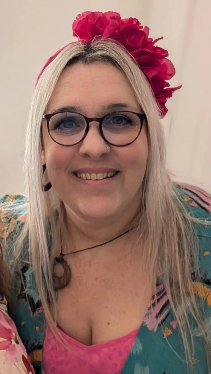 Women with blonde hair wearing glasses, a pink flower headband, and a colorful floral dress, smiling at the camera.