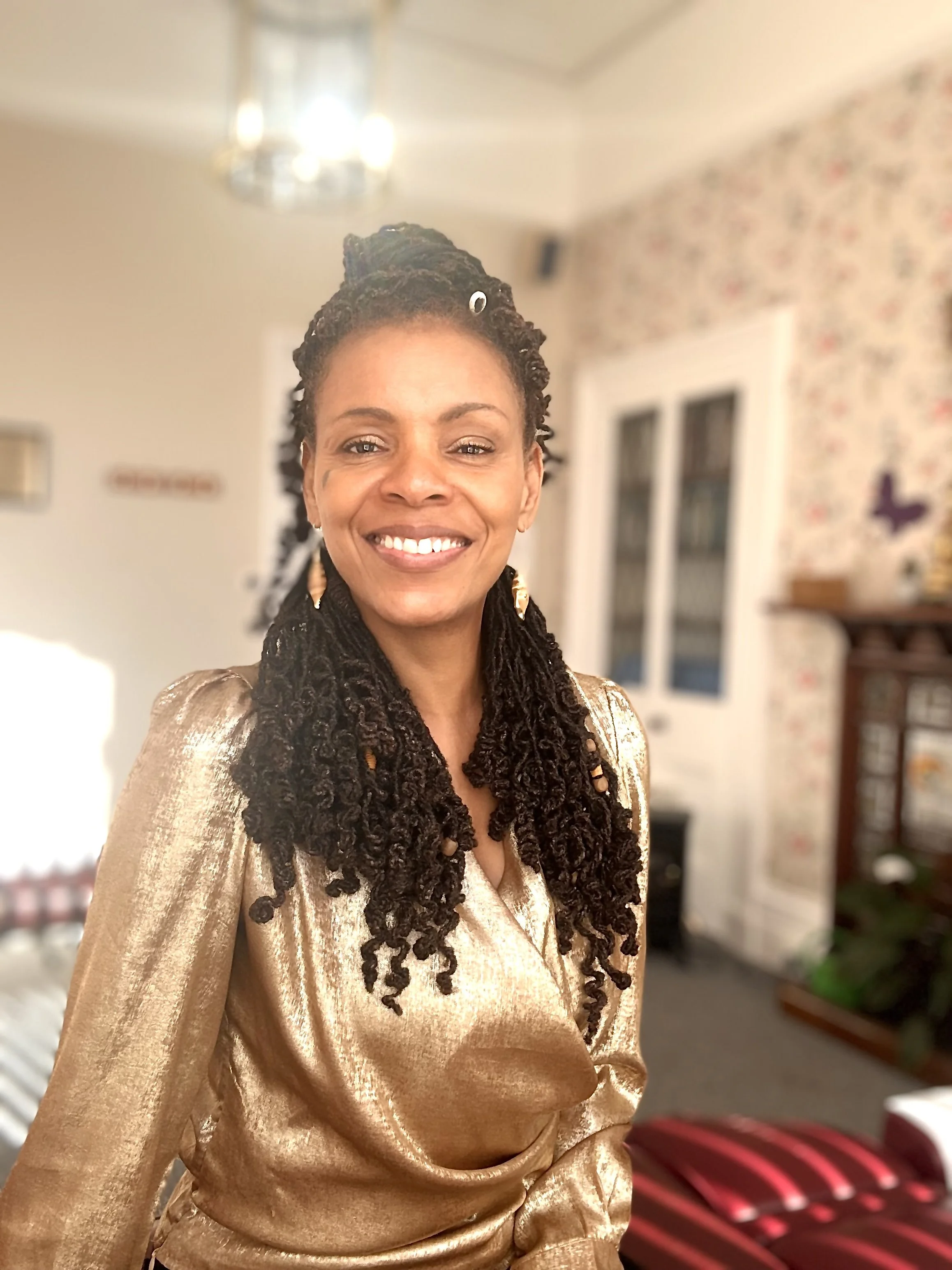 A smiling woman with natural braided hair, wearing a shiny gold dress, standing in a warmly lit living room with floral wallpaper and furniture.