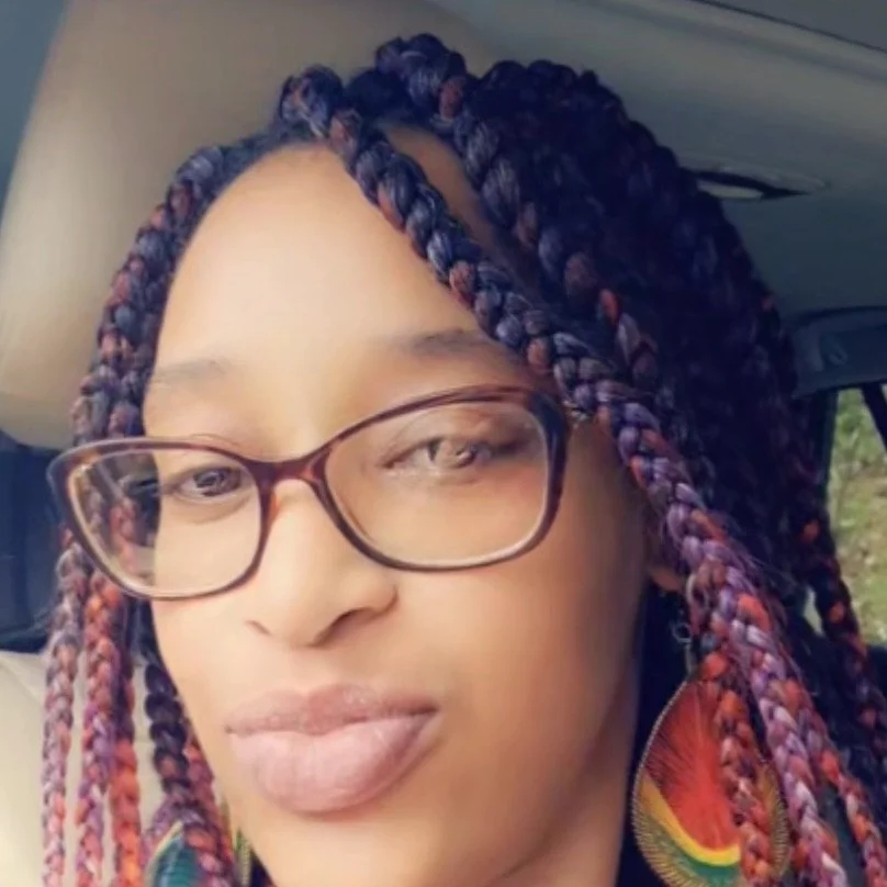 Close-up of a woman with colorful braided hair, glasses, and earrings, sitting in a car.