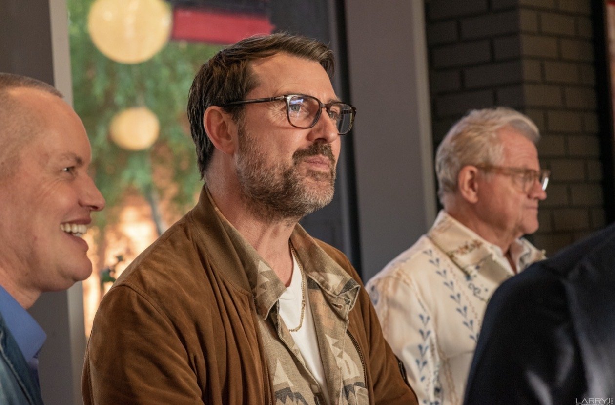 Three men sitting indoors at an event, with the man in the middle having dark hair, glasses, a beard, and wearing a brown jacket. The man on the left is smiling, and the man on the right has gray hair and glasses, wearing a patterned shirt.