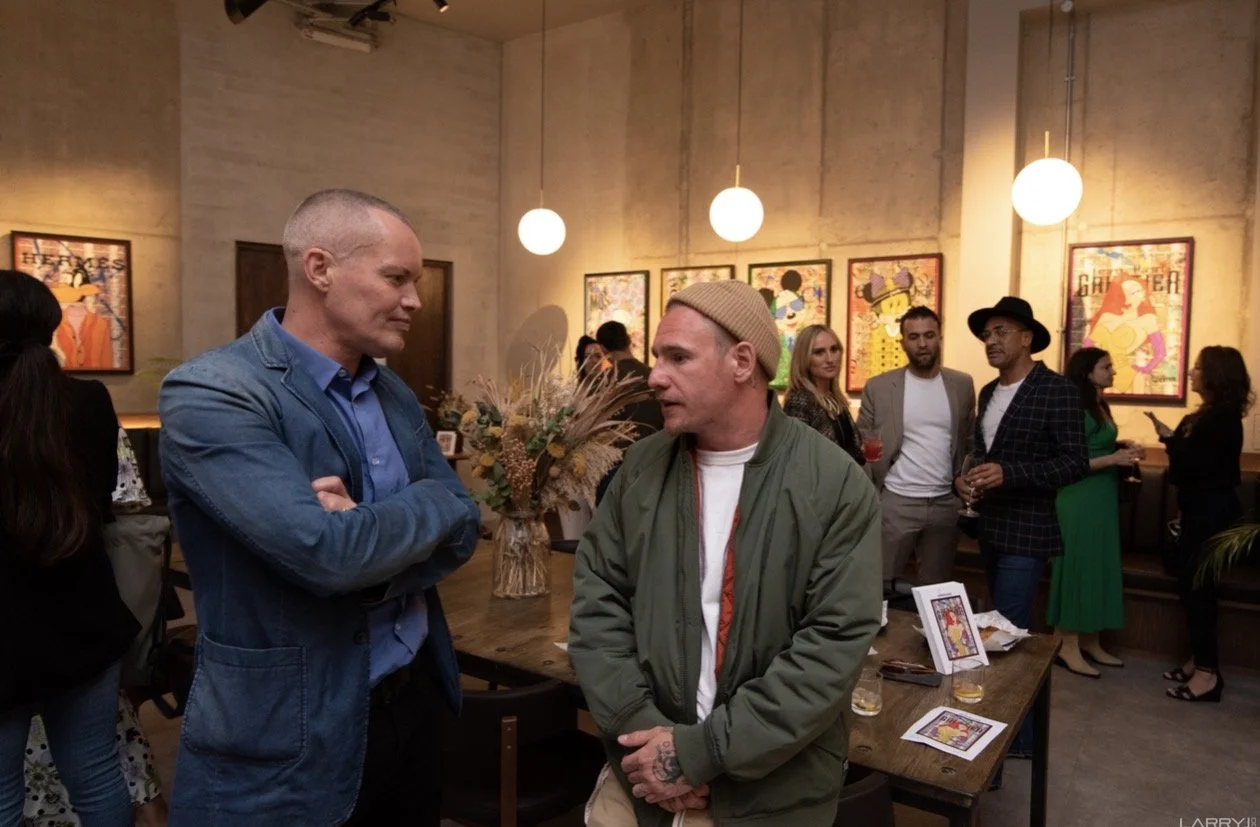 A social gathering indoors with several people engaged in conversations. Two men are in the foreground, one with a shaved head wearing a blue jacket with arms crossed, and the other wearing a beige beanie and green jacket, talking to each other. The 