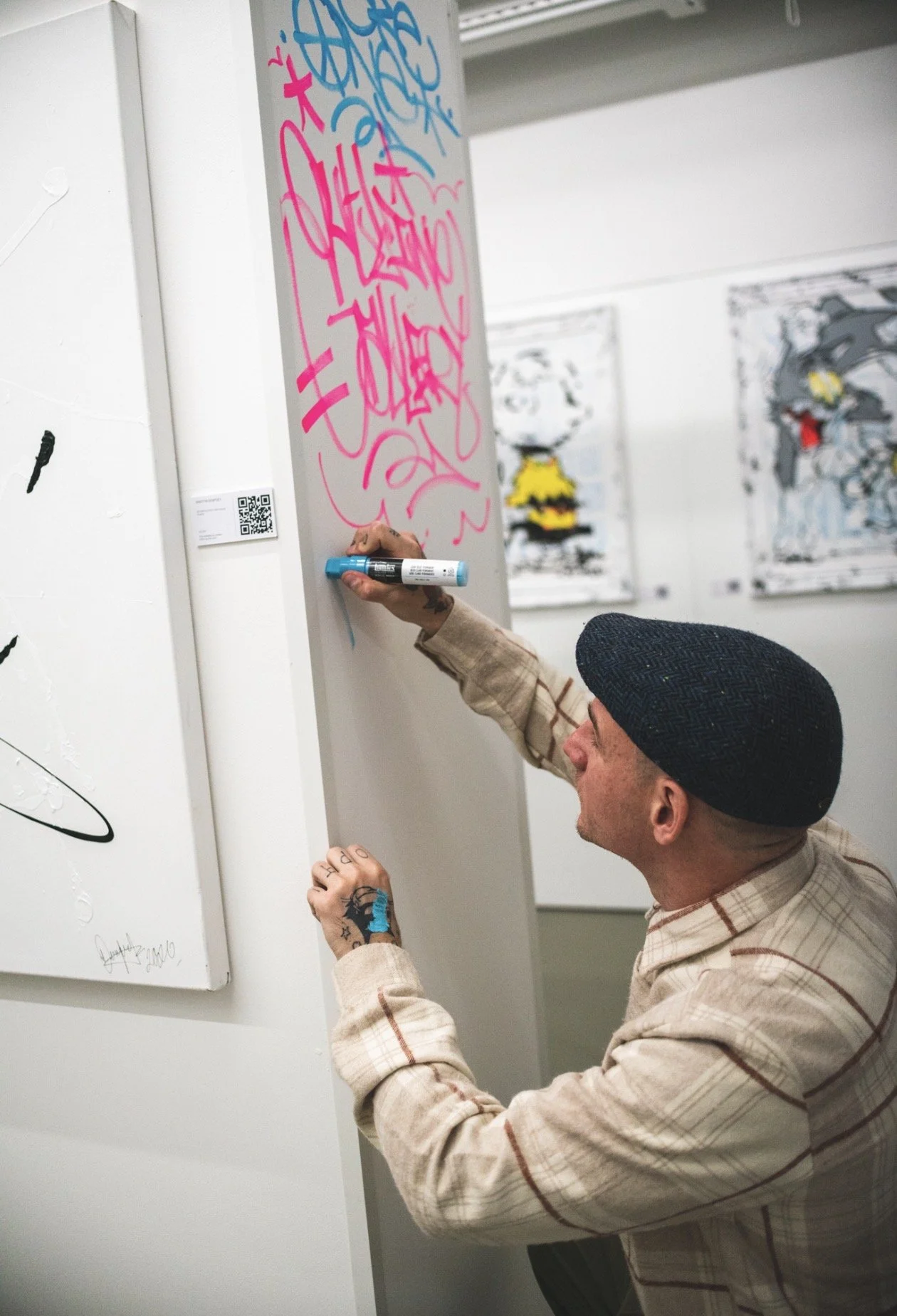 An artist wearing a black cap and a beige plaid shirt is drawing on a vertical surface with a pink marker, creating graffiti-style art. The background shows a white wall with various artworks displayed.