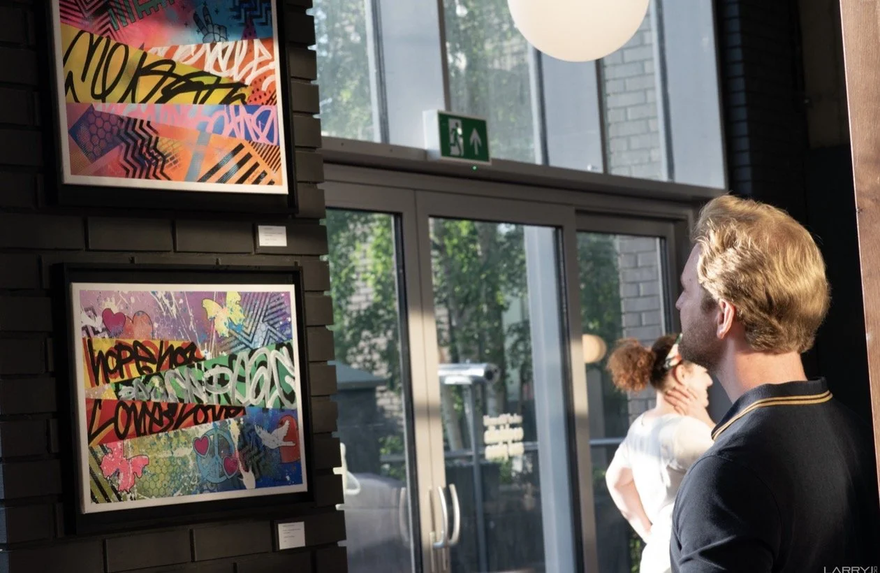 People viewing colorful graffiti-style artwork on a black brick wall in a gallery or exhibition space with large windows and a green emergency exit sign.