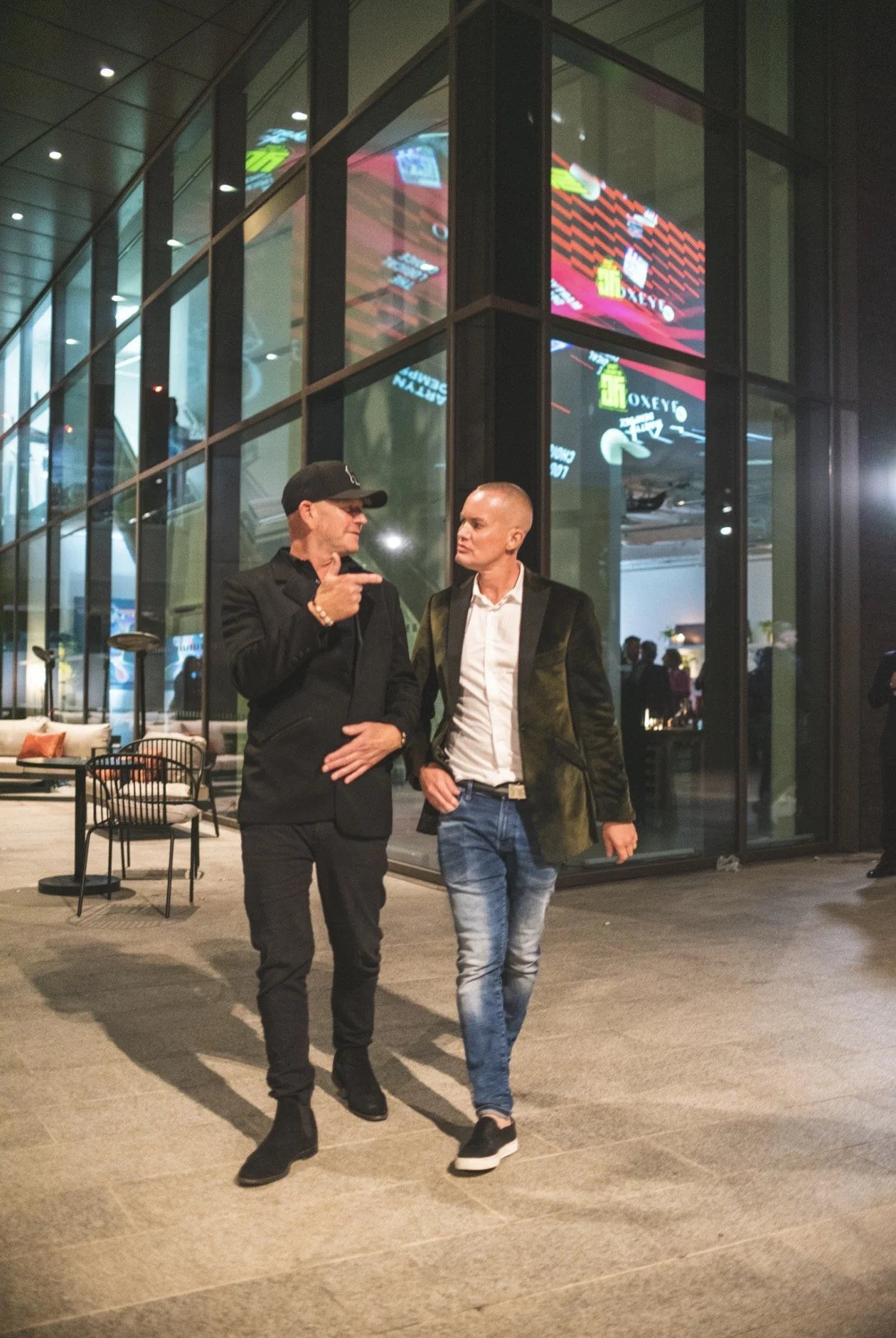 Two people, a man and a woman, walking and talking outside a modern building with large glass windows at night.