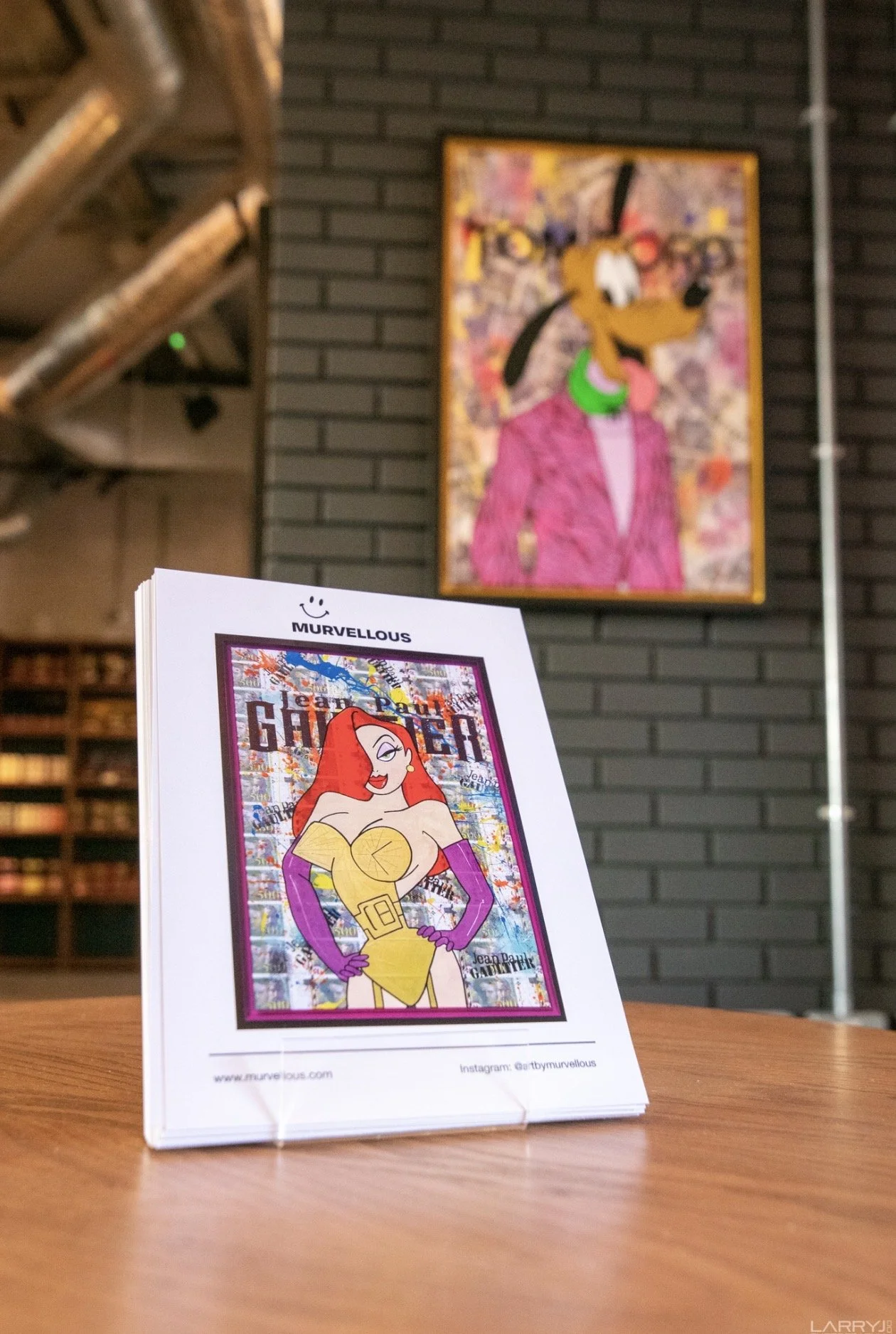 Table tent displaying a cartoon drawing of a female character with long red hair, purple gloves, and a yellow dress, titled "GAMER" with graffiti background; in the background, a colorful portrait of a cartoon animal with glasses on a dark wall.