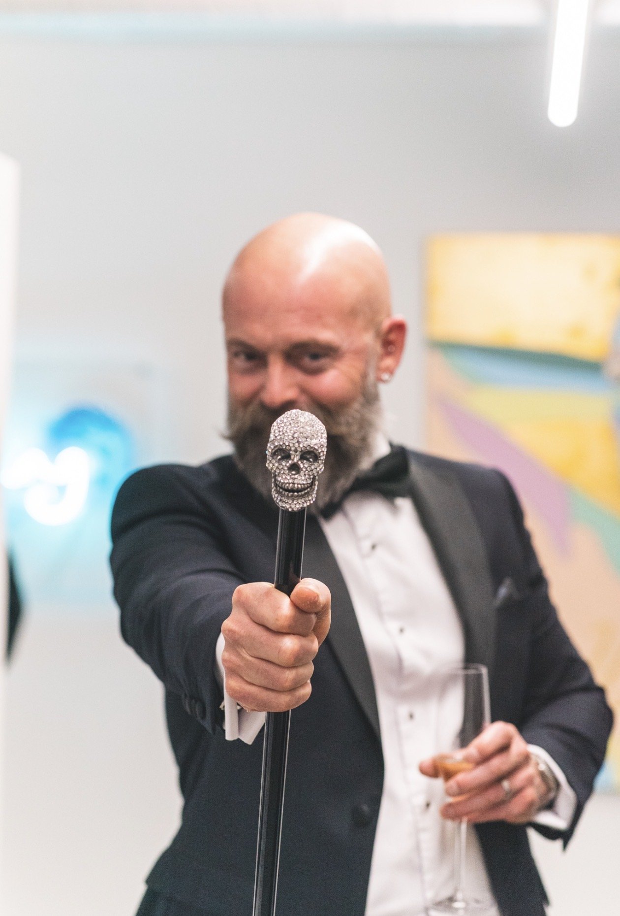A man in a tuxedo holding a skull-shaped decorated cane and a glass of champagne at a celebration or event.