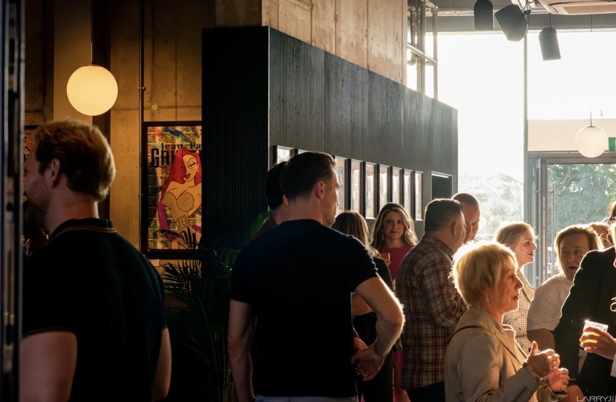 People gathering in a modern indoor space with warm lighting, artwork on the walls, and large windows letting in natural light.