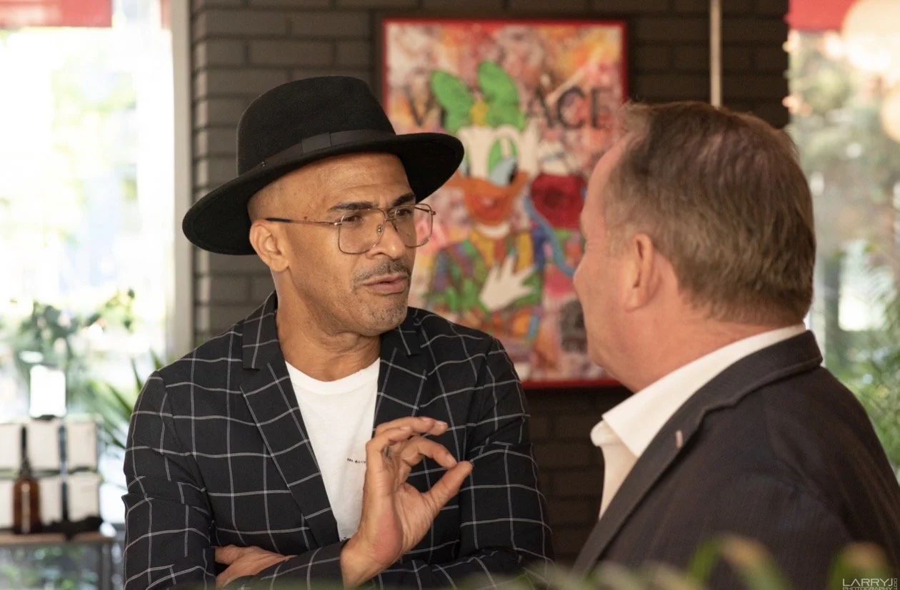 Two men engaged in a serious conversation indoors, one wearing glasses, a black hat, and a black and white checkered blazer, and the other with short hair and a dark suit. In the background, there is colorful artwork on the wall and large windows all