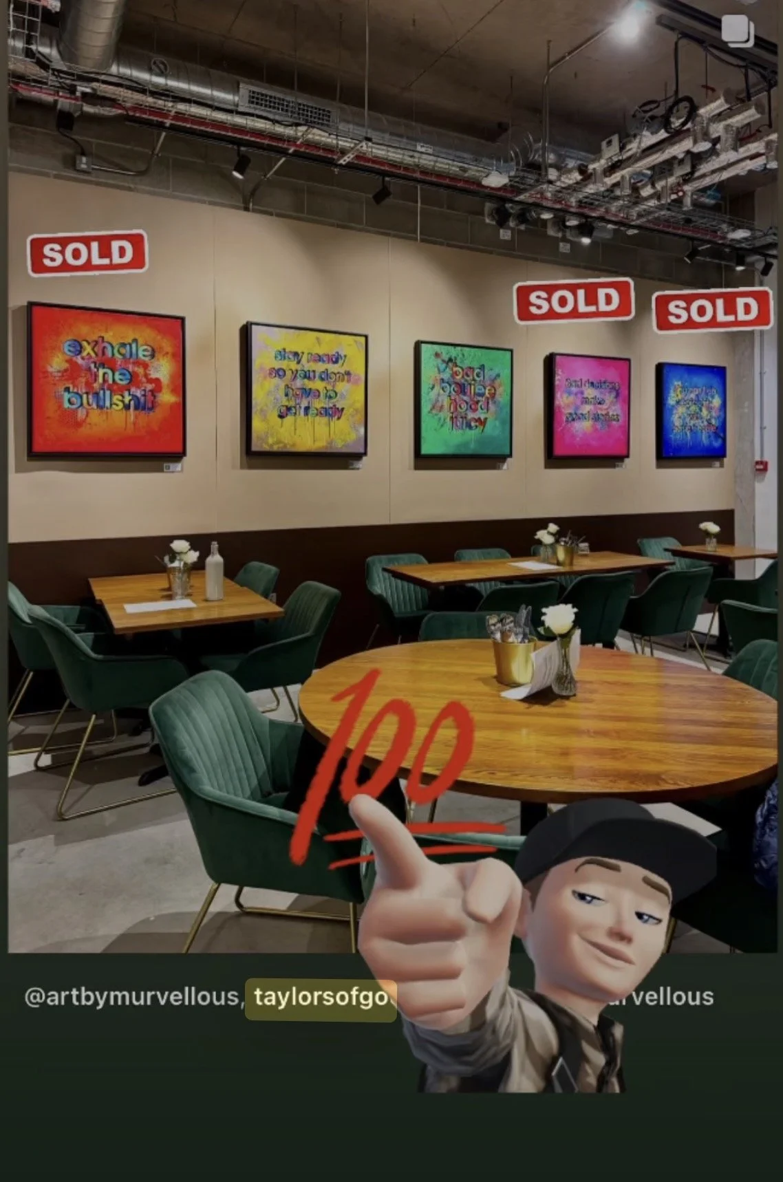 A cozy cafe interior with wooden tables and green chairs. The wall features colorful framed art with funny phrases and several are marked with 'SOLD' signs. A cartoon character with a pointing finger drawing and the number 100 written in red appears 