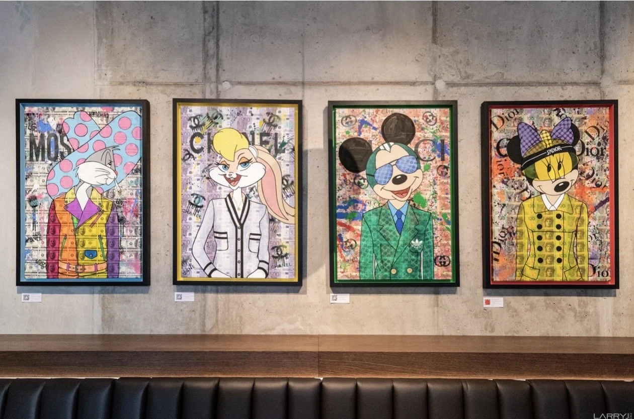 Four colorful framed artworks of cartoon characters on a concrete wall in an art gallery, with a black leather bench in front.