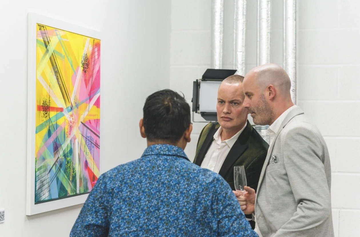 Three men in conversation at an art gallery, with colorful abstract artwork on the wall.