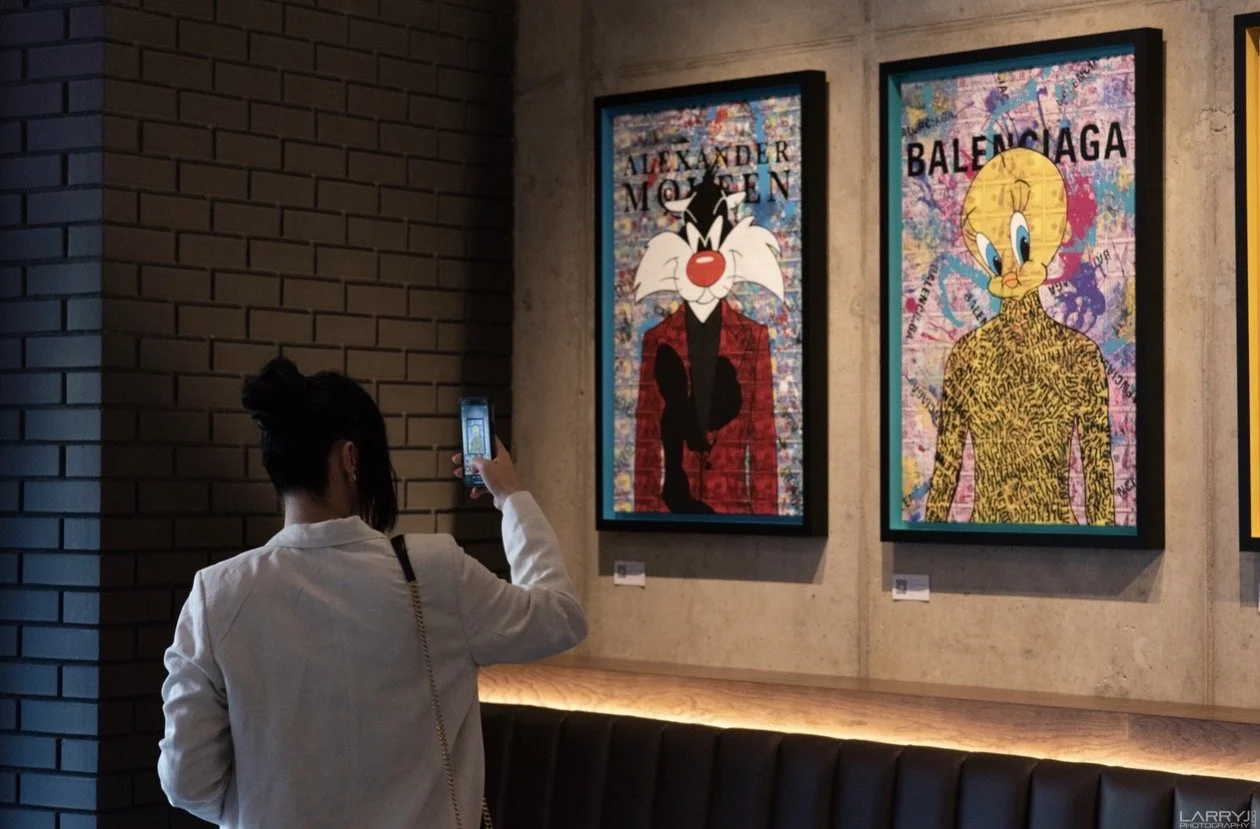 A woman in a white blazer is taking a photo with her phone of two framed colorful pop art paintings on a wall. 