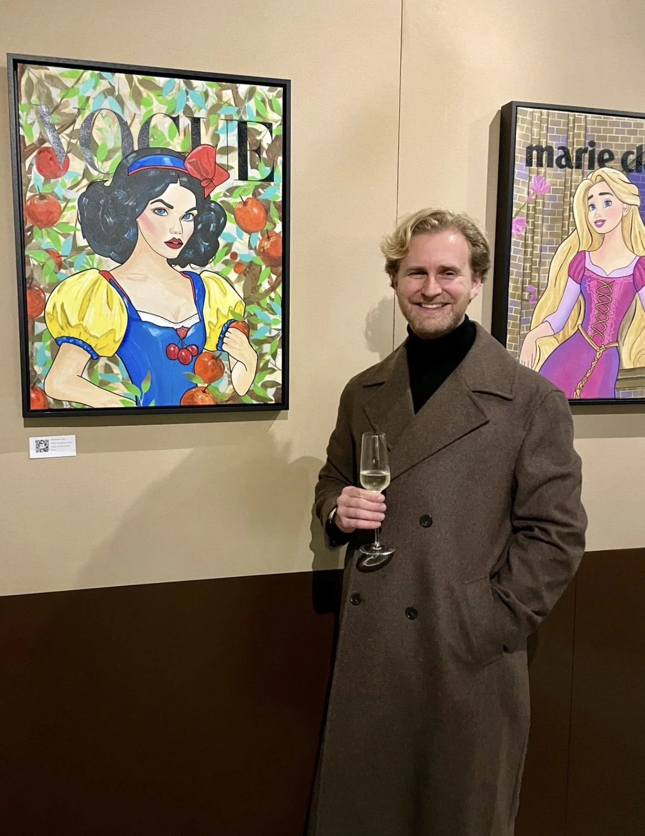 A man in a brown coat holding a glass of champagne standing in front of colorful paintings of  princesses at an art gallery.