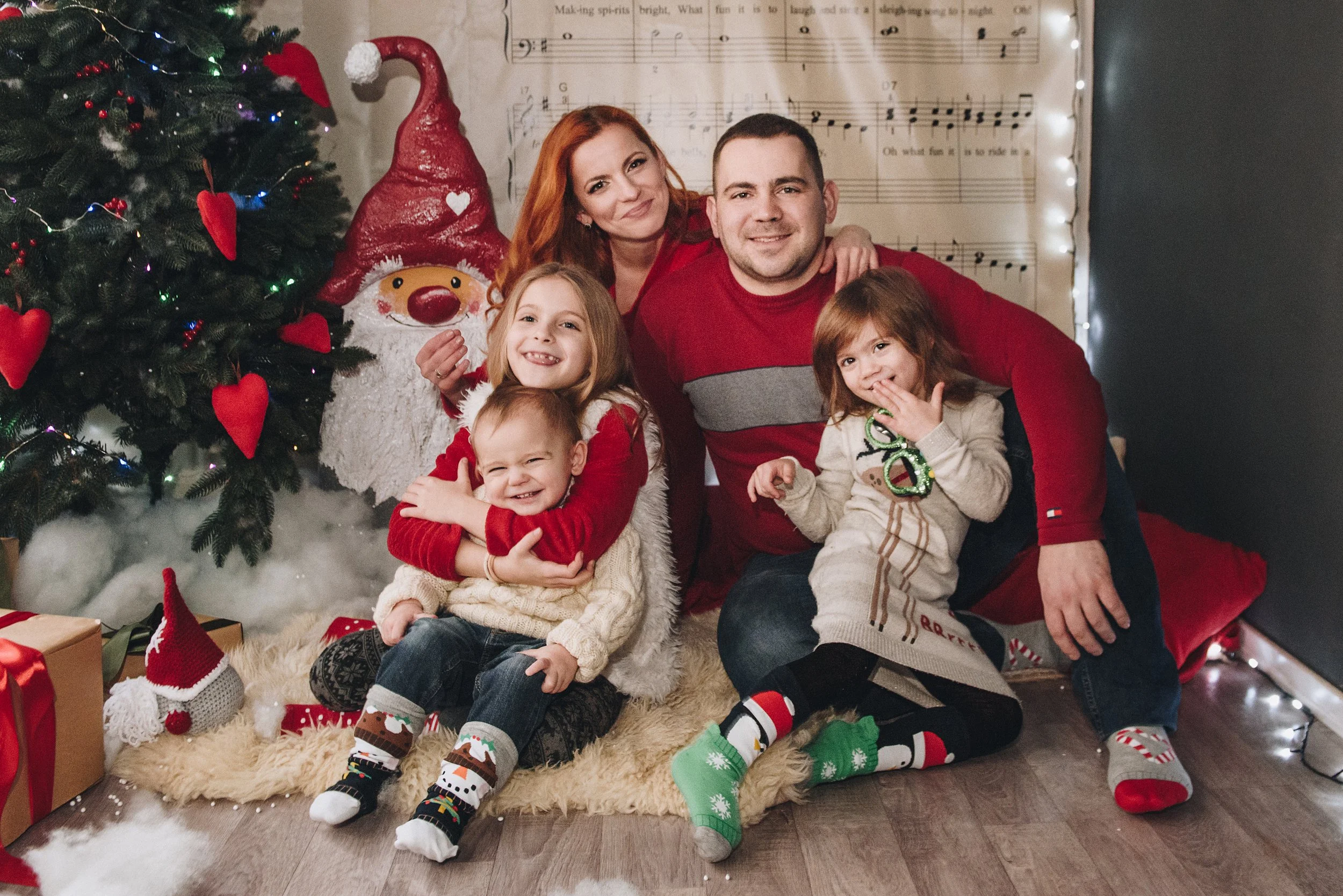 A family of five smiling and sitting on a furry rug in front of a decorated Christmas tree. The family includes a woman, a man, and three young children. The scene is festive with Christmas stockings, wrapped presents, and holiday decorations.