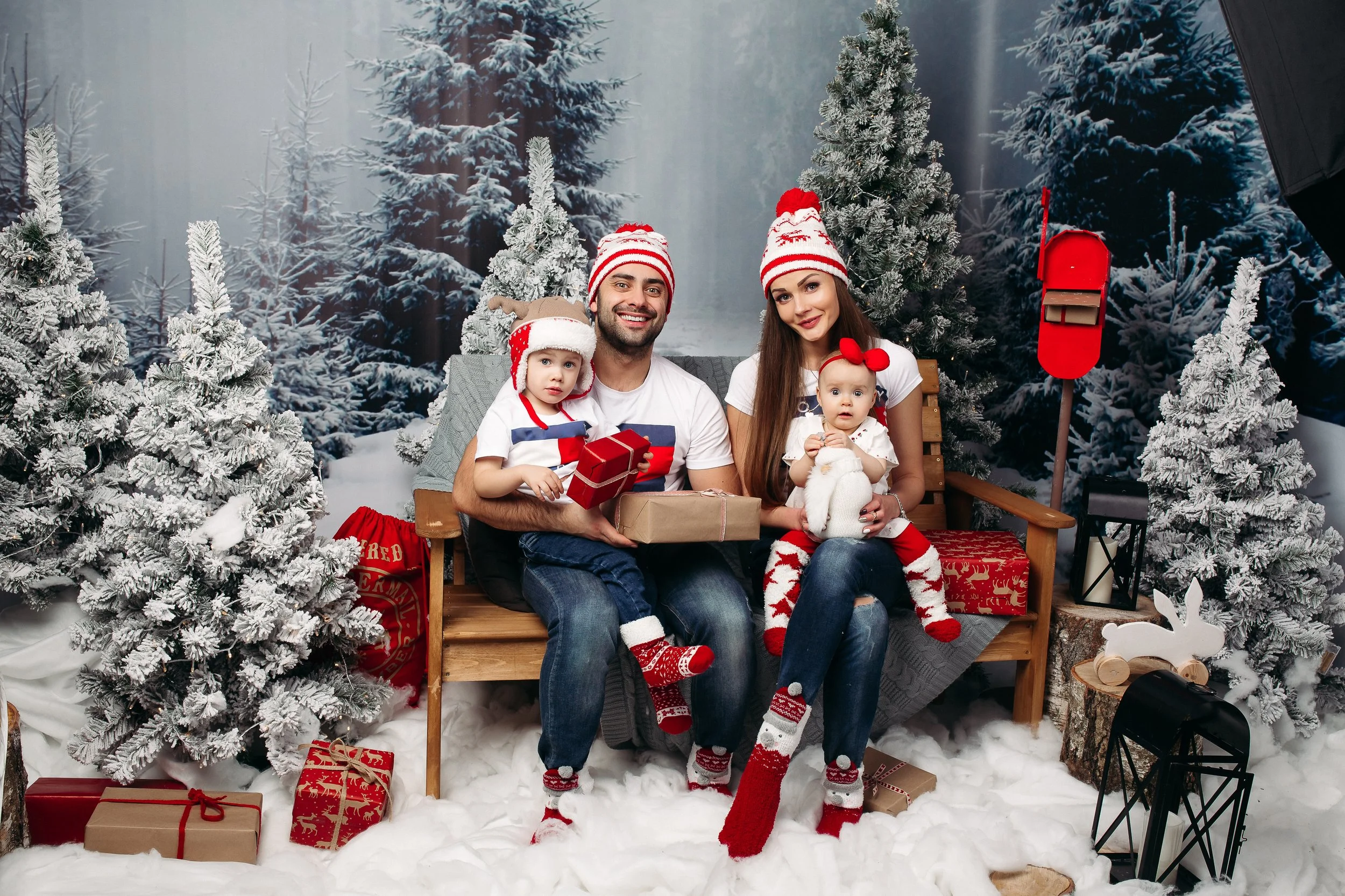 Family sitting on a wooden bench in a snowy Christmas scene, surrounded by snow-covered trees, with gifts, and holiday decorations, wearing festive hats and socks.