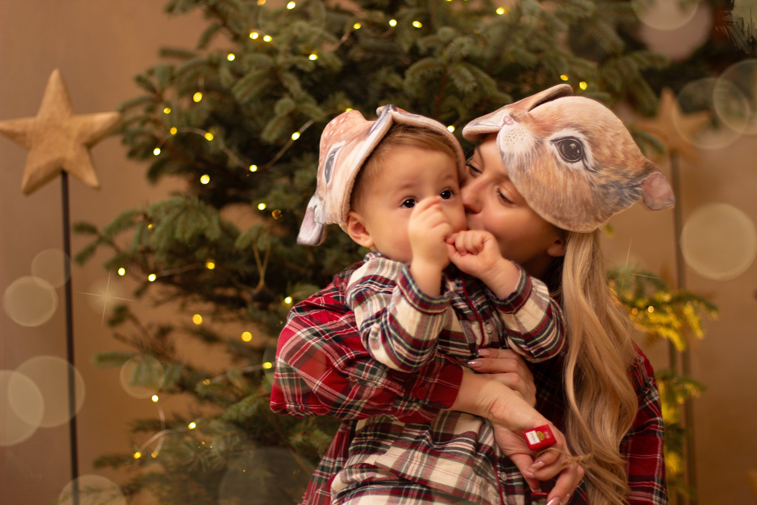 A woman and a young child wearing animal-themed hats, with the woman kissing the child on the cheek in front of a decorated Christmas tree.