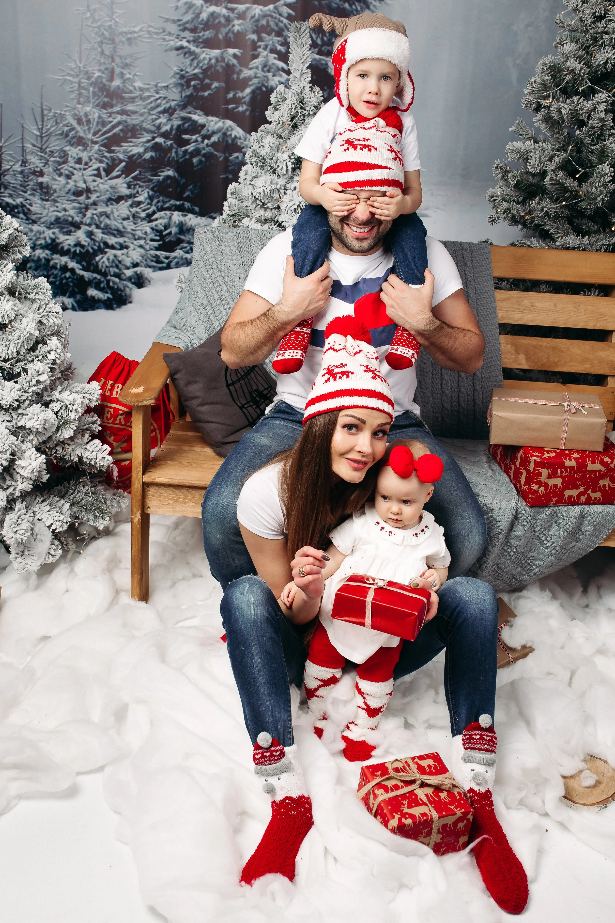 Family of four dressed in Christmas attire, sitting on a snowy floor with Christmas trees and presents, celebrating Christmas.
