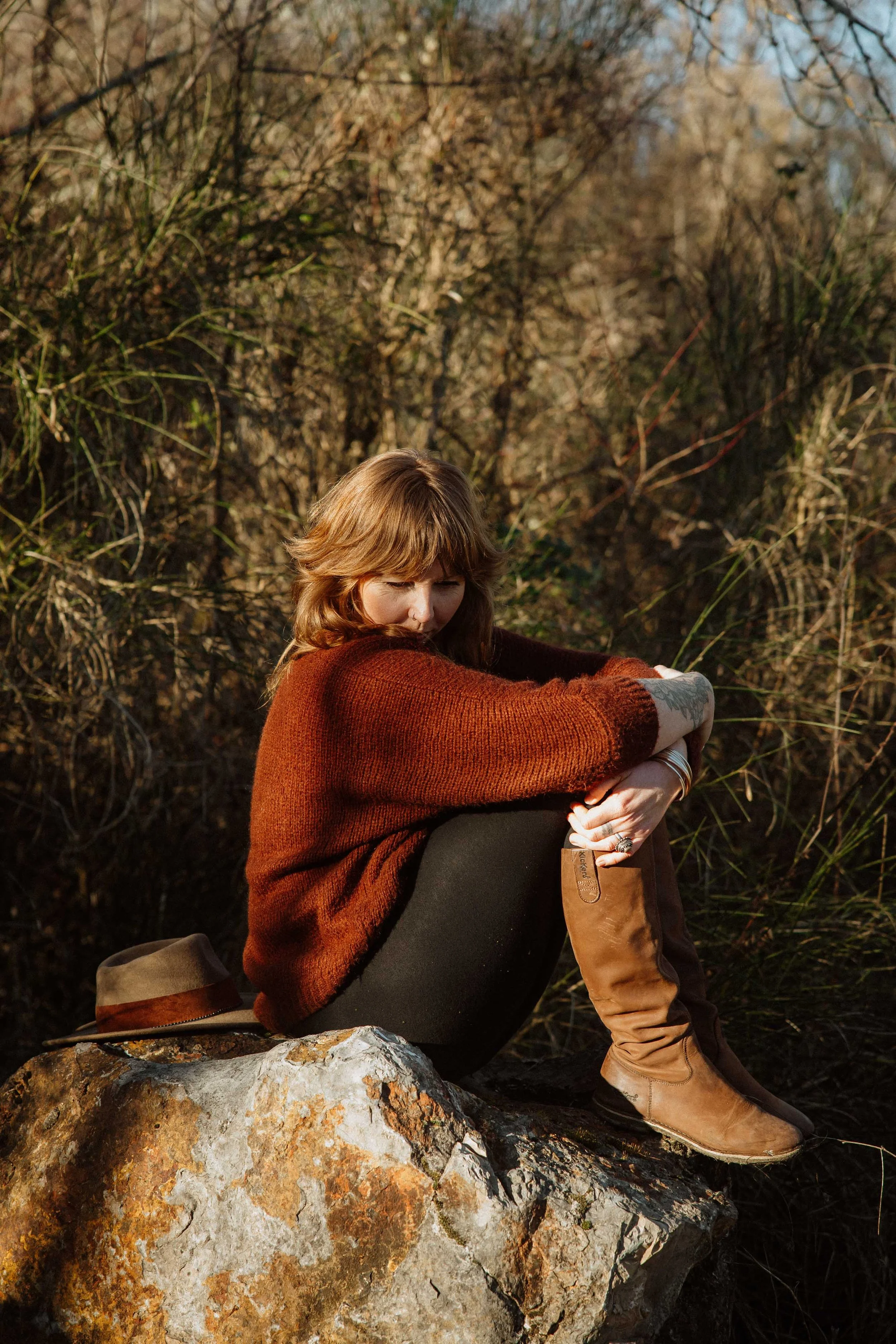 Femme assise sur une roche dans la nature, portant un pull marron foncé et des bottes en cuir, avec un chapeau posé à côté.