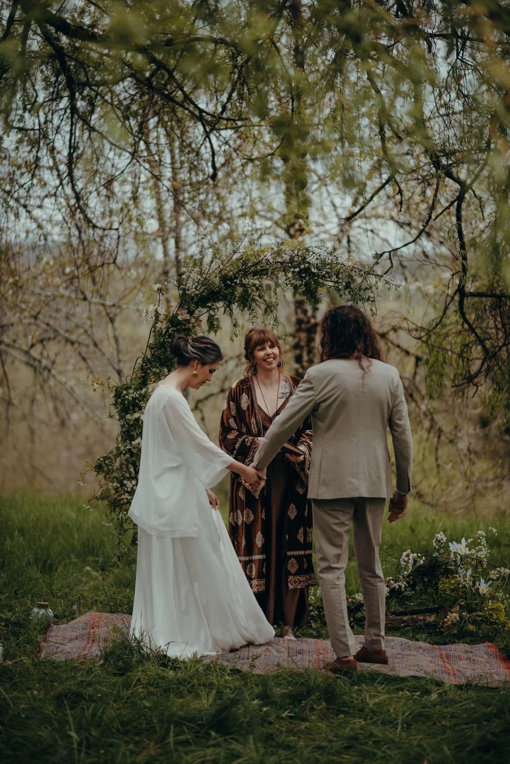 Un couple se marie lors d'une cérémonie en plein air, devant une officiante, sous un arc floral, dans un environnement naturel avec des arbres en arrière-plan. Mariage Hautes-Pyrénées. Mariage Bagnère