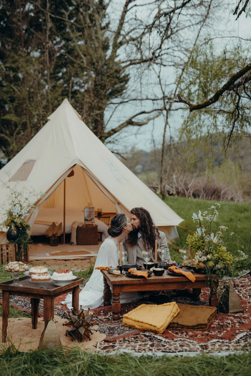 Un couple s'embrasse le jour de leur mariage écolo devant une tente en plein air, avec une table de pique-nique décorée de fleurs, de nourriture et de boissons, lors d'un pique-nique romantique en nature.