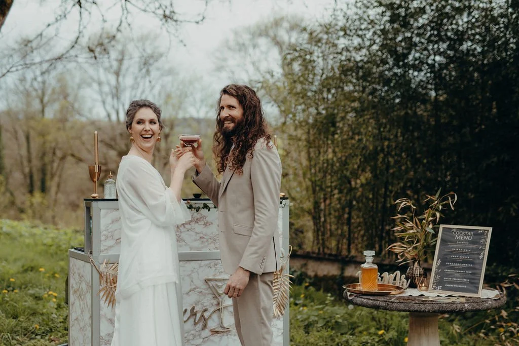 Cérémonie de mariage holistique et alternatif célébrée par Judith La Dame de Cœur : un couple souriant tenant un verre à cocktail dans un décor extérieur avec bar et menu de cocktails sur la table