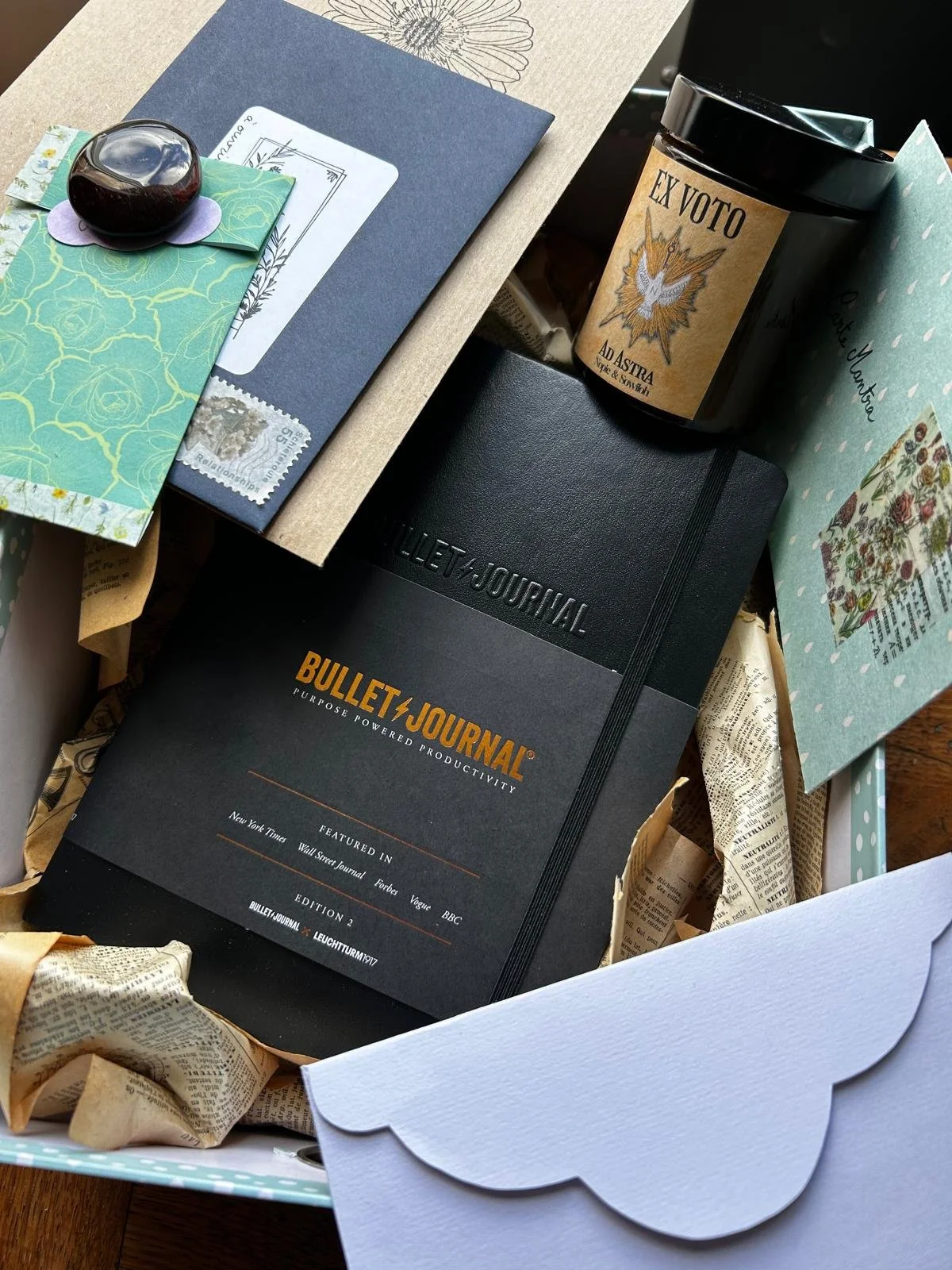 Une boîte contenant un carnet Bullet Journal noir, une bougie parfumée avec une étiquette "Ex Voto", plusieurs enveloppes ou cartes décoratives avec des motifs floraux et une fenêtre en forme de nuage, ainsi que du papier de saison Kit art-coaching