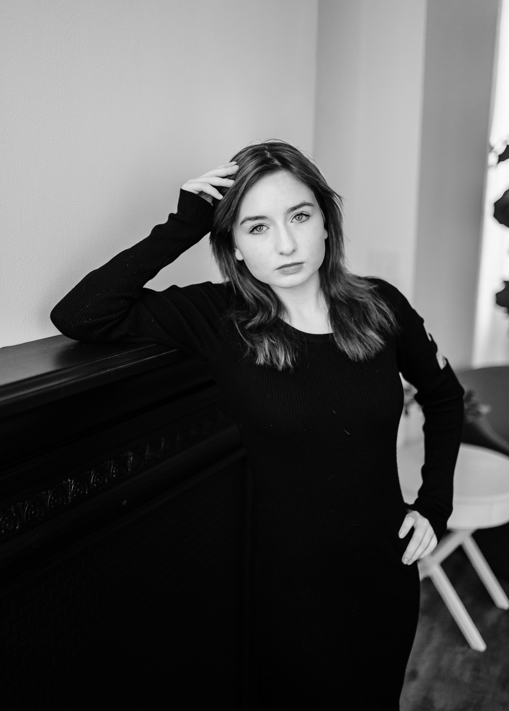 A young woman with light skin and medium-length hair, wearing a black long-sleeve top, leaning against a dark wooden piece of furniture in a room with light-colored walls. She has one hand on her hip and the other resting on her head, looking directl