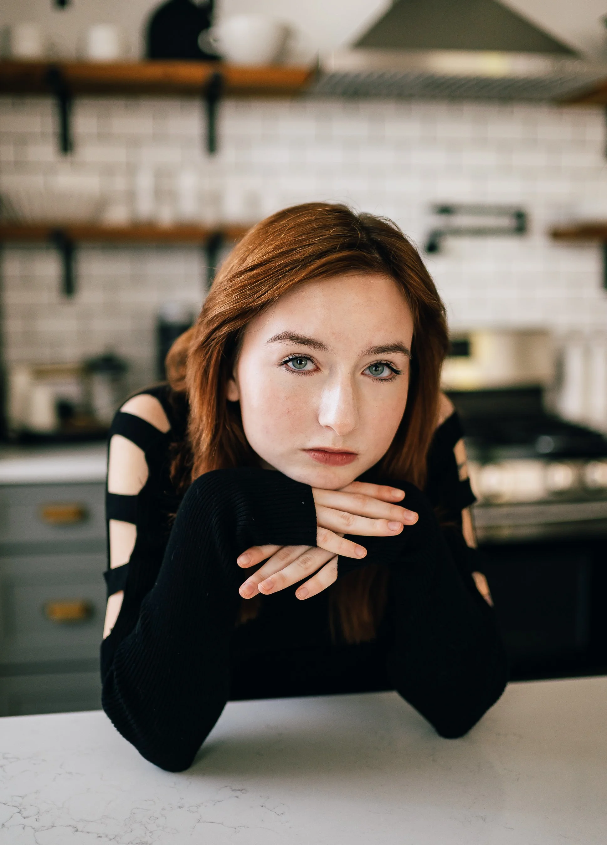A young woman with red hair and blue eyes resting her chin on her hands at a kitchen counter, wearing a black sweater with cut-out shoulder details.