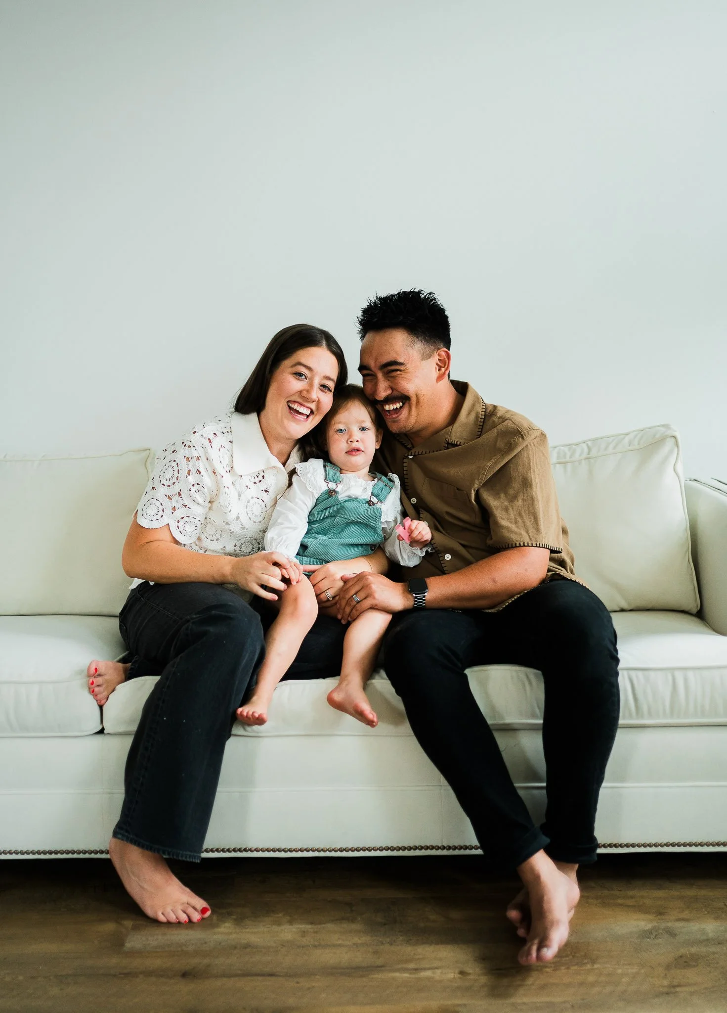 A family of three sitting on a cream-colored sofa, smiling and enjoying time together. The woman has short brown hair and is wearing a white eyelet top. The man has short dark hair and is wearing a tan shirt. Their young daughter, with light brown ha