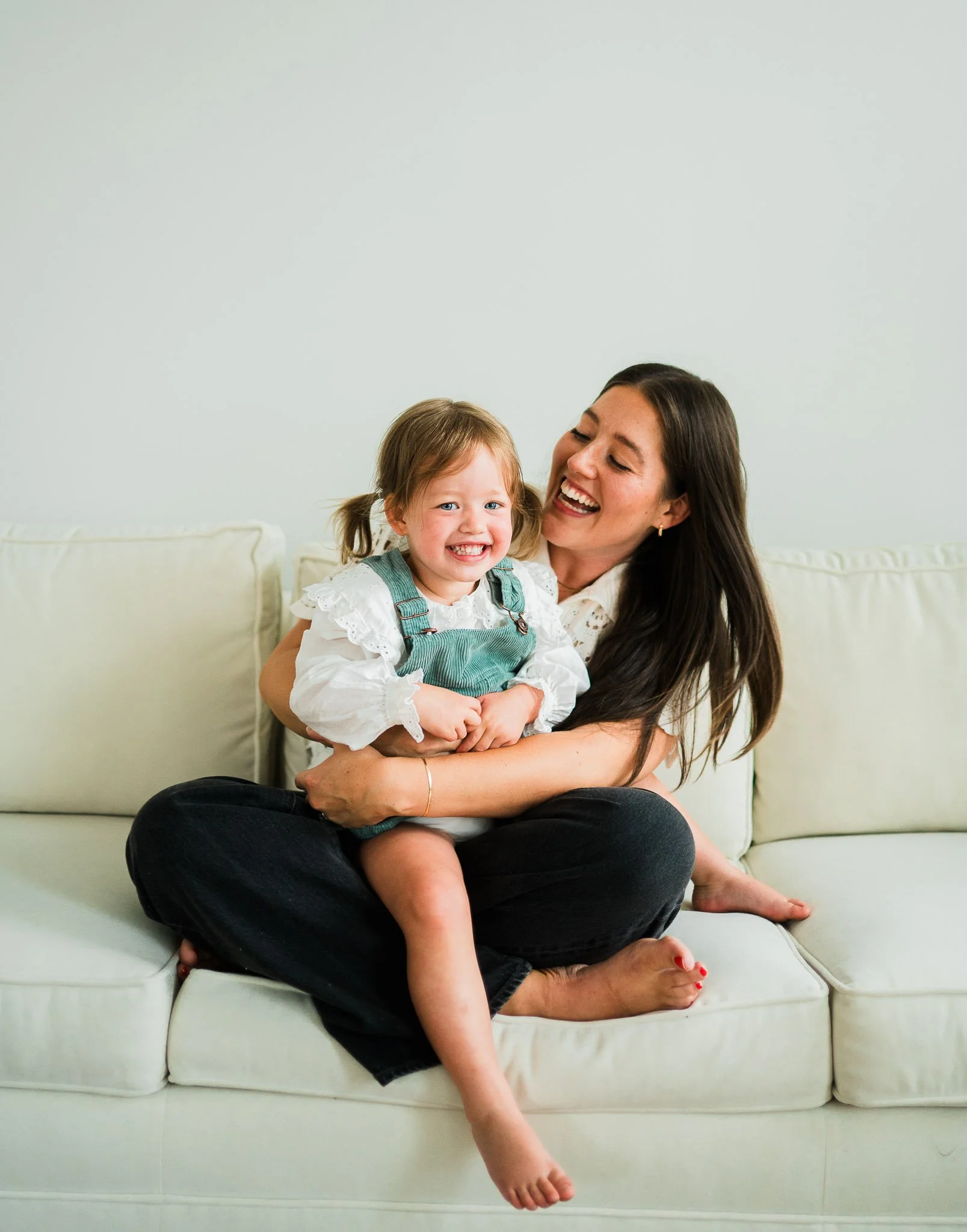 A woman and a young girl sitting on a white sofa, smiling and embracing each other. The woman has long dark hair and the girl has light brown hair in pigtails.