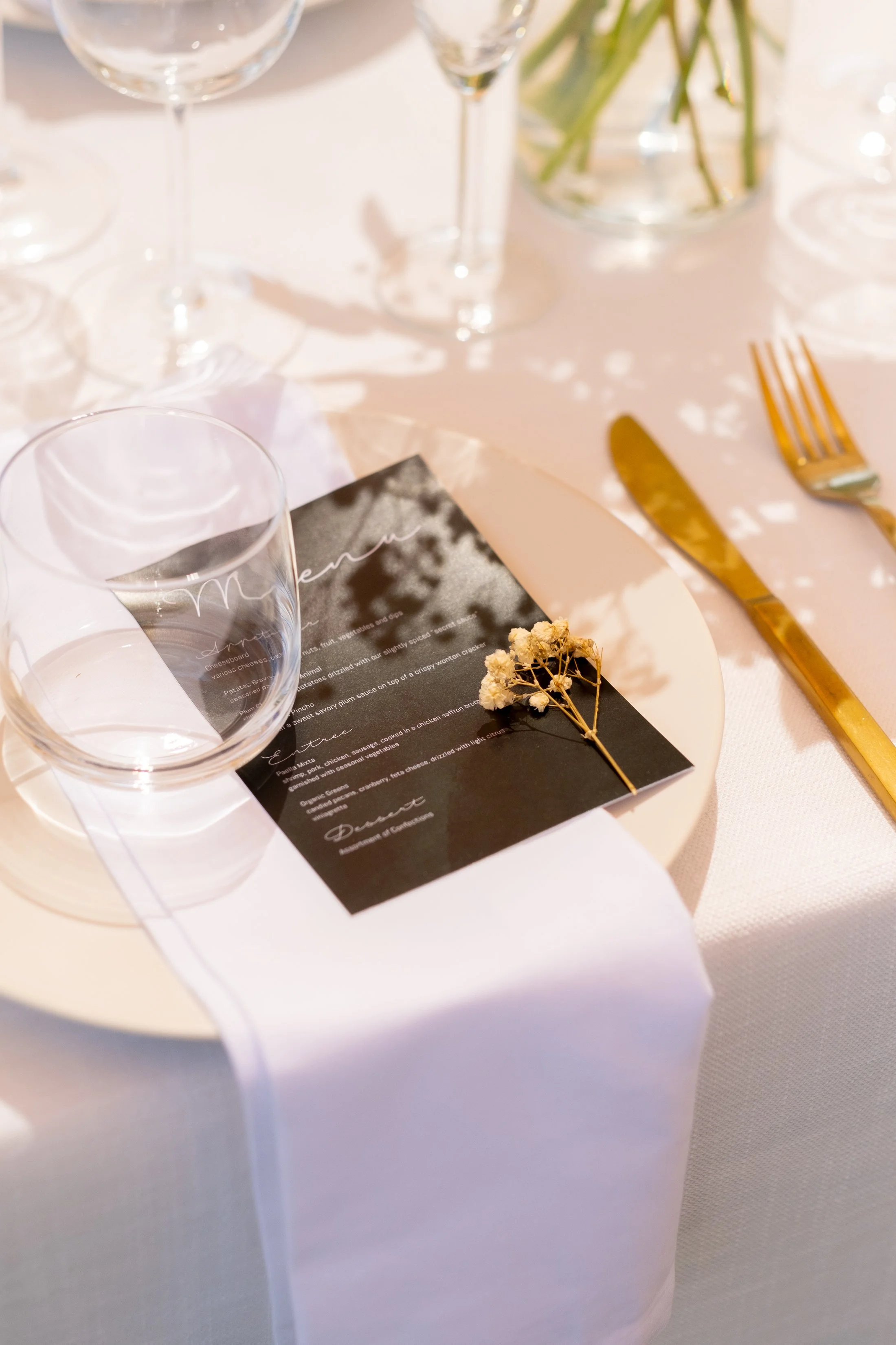 A formal table setting with wine glasses, a black menu card, a white plate with a white tablecloth, gold utensils, and a flower in a vase, with a sprig of dried flowers on top of the menu.