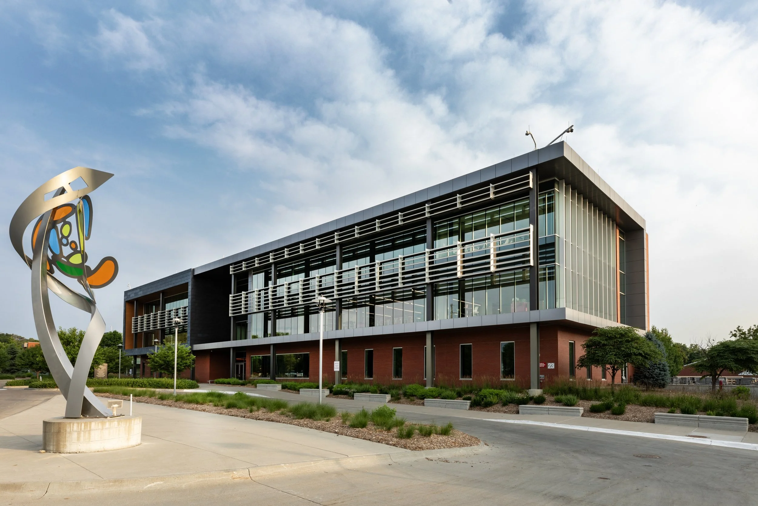 A modern building with large glass windows and a metal sculpture in front. The structure features brick and steel elements, and the surrounding area has landscaped greenery and a paved entrance.