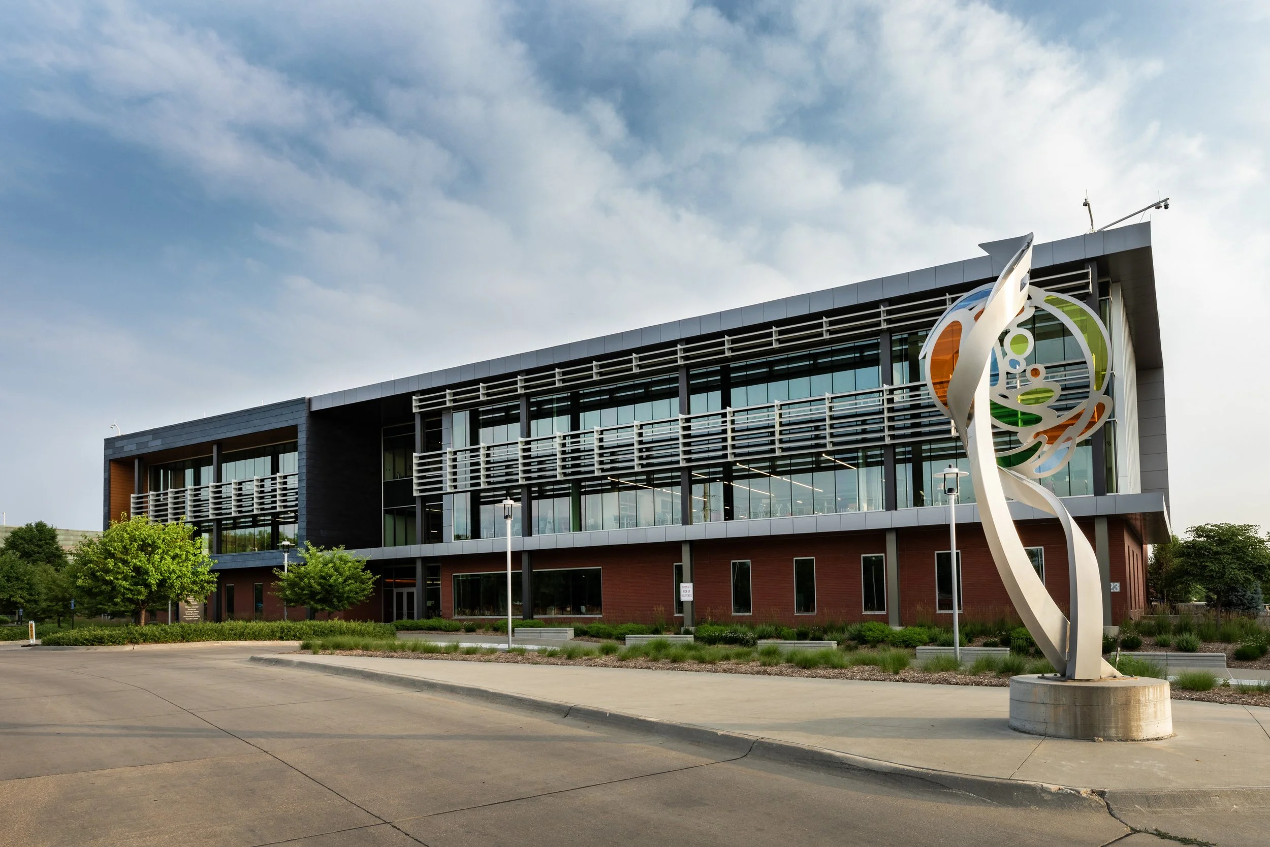 Modern office building with large windows and metal accents, featuring a colorful abstract sculpture in the courtyard.