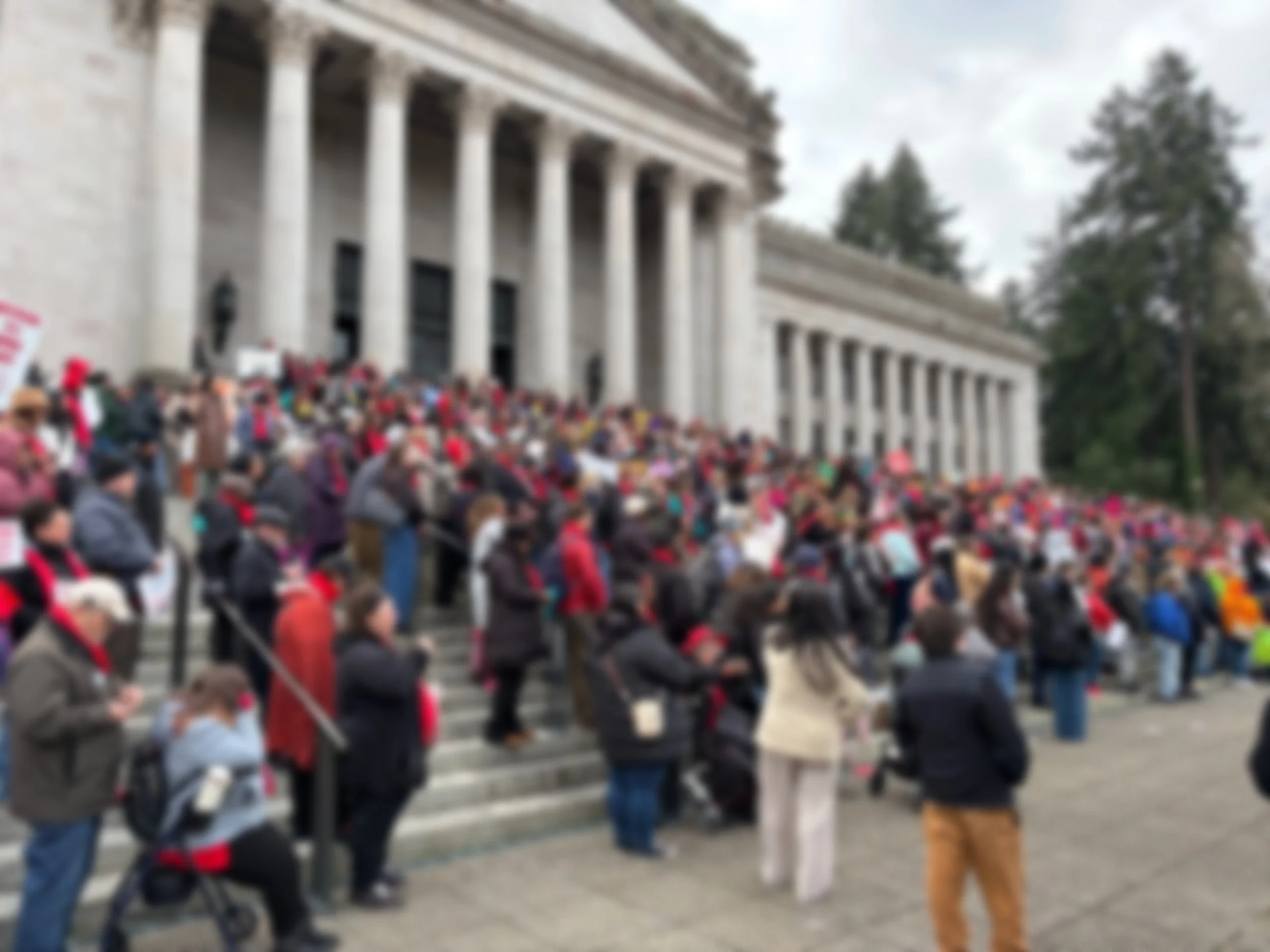 Large group of advocates gathered on the steps of a government building with tall columns, holding signs advocating for rent caps and housing rights.