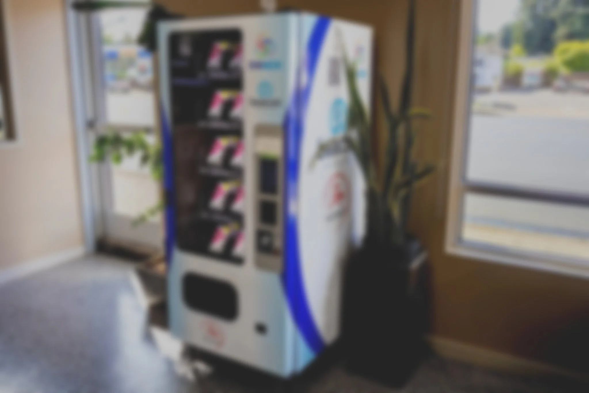 Vending machine next to a window with outside view and potted plant.