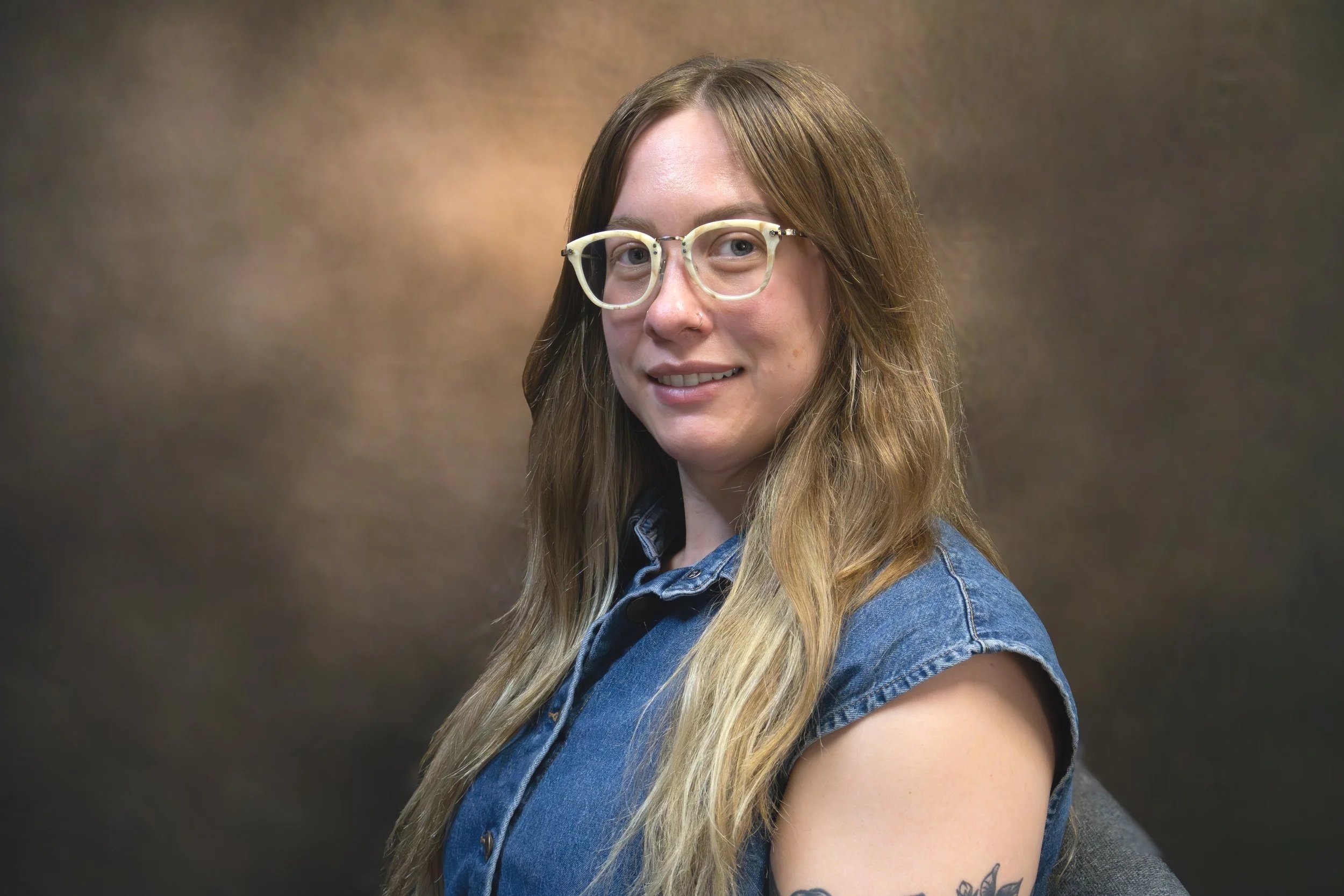 A woman with long wavy blonde hair, wearing glasses and a white button-up shirt, smiling at the camera against a gray backdrop.