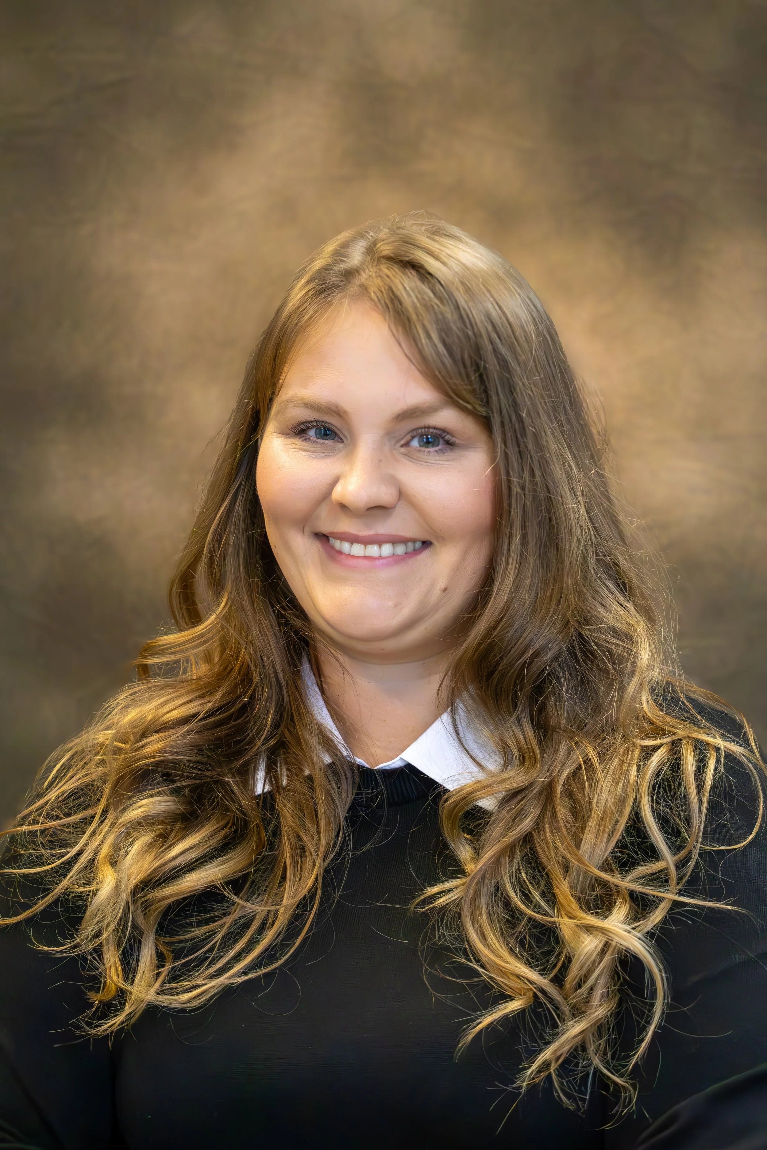 Portrait of a smiling young woman with long, wavy, light brown hair, wearing a black top over a white collared shirt, against a textured brown background.