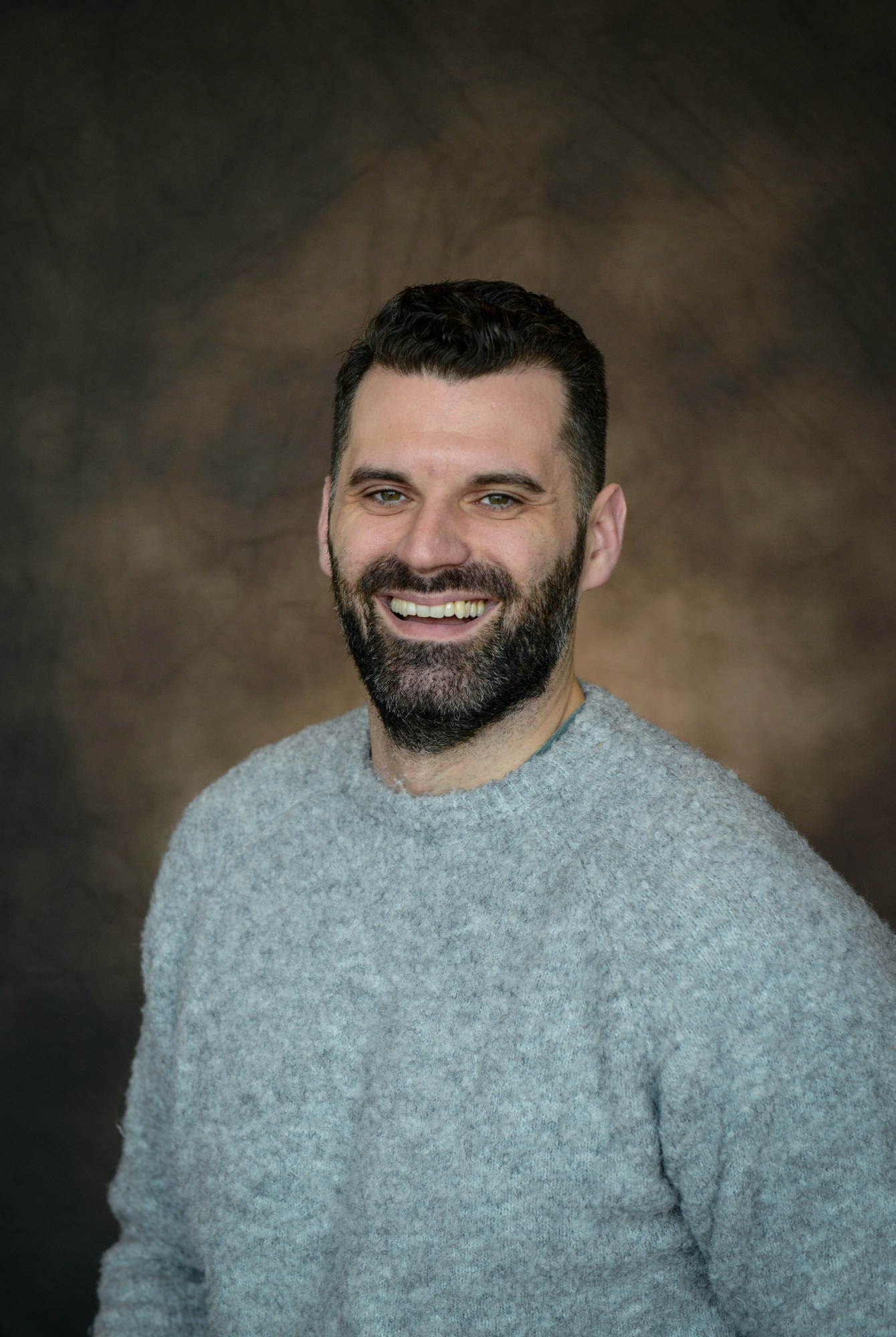 A smiling man with dark hair, a full beard, and wearing a dark gray button-up shirt standing in front of a gray textured background.
