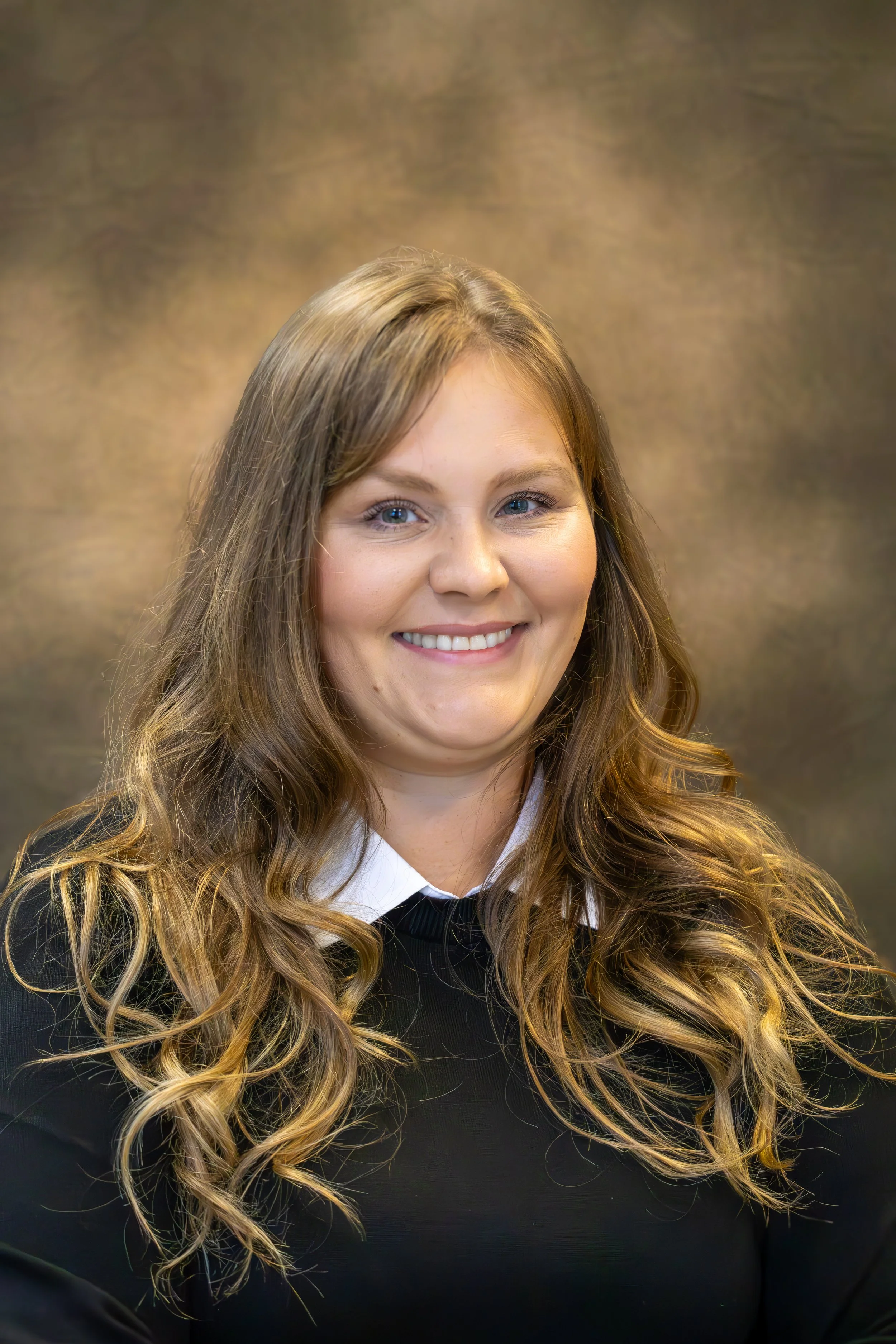 Portrait of a smiling young woman with long, wavy, light brown hair, wearing a black top over a white collared shirt, against a textured brown background.
