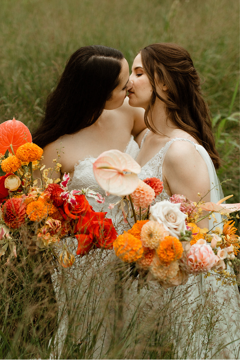 Two brides kiss while wearing white dresses and holding red and orange flowers at a Minneapolis wedding