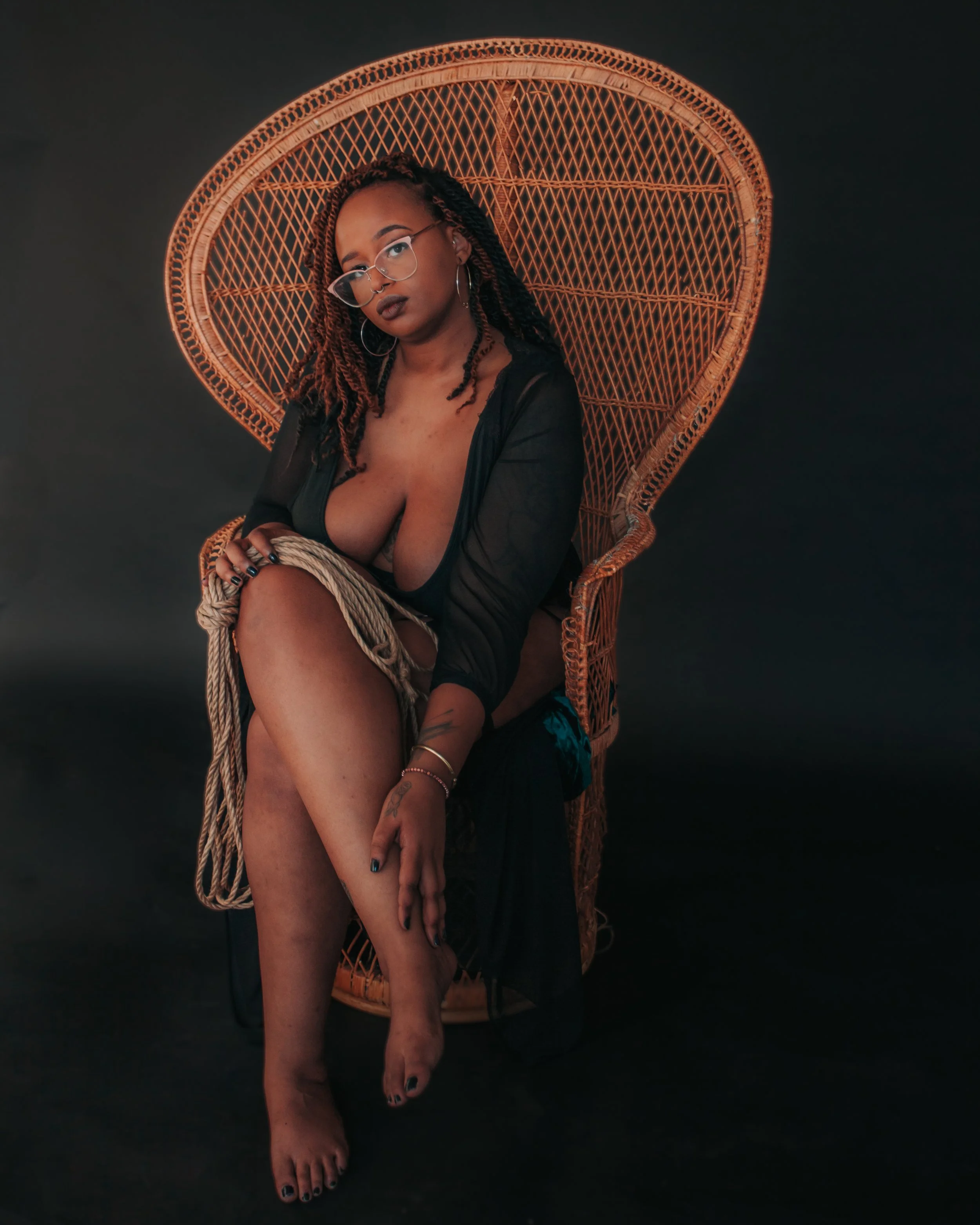 Woman with braided hair and glasses sitting on a wicker chair against a dark background, wearing a black sheer dress and jewelry.