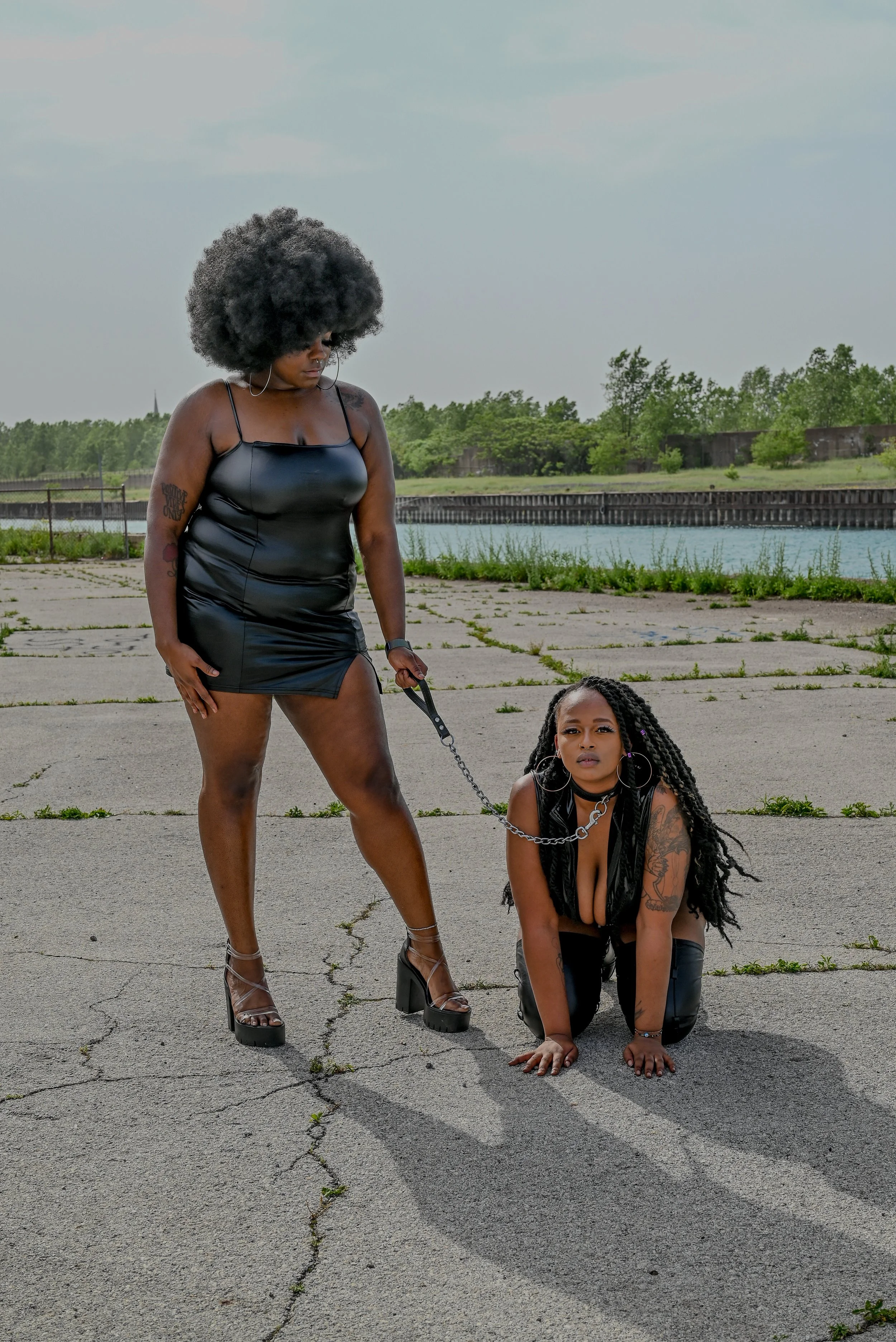 Two women with tattoos and dark clothing on cracked pavement near water. One woman with curly afro hair stands holding a chain connected to the other woman, who is kneeling on the ground with long braids and tattoos, looking directly at the camera.