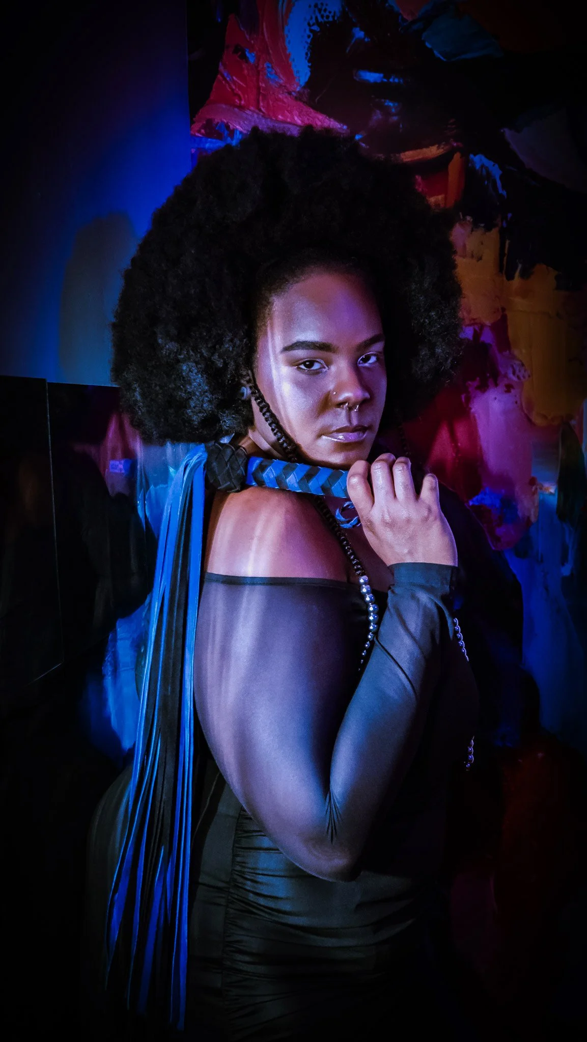 A young woman with natural curly hair, wearing a black off-shoulder dress with sheer long sleeves, holding a blue patterned tie over her shoulder, standing against a colorful abstract background with dark blue, pink, and red tones.